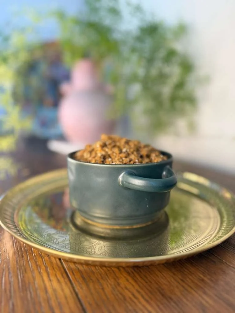 A small blue ceramic cup filled with panjiri on a gold decorative plate on a wooden table, with a blurred background of plants and vases.