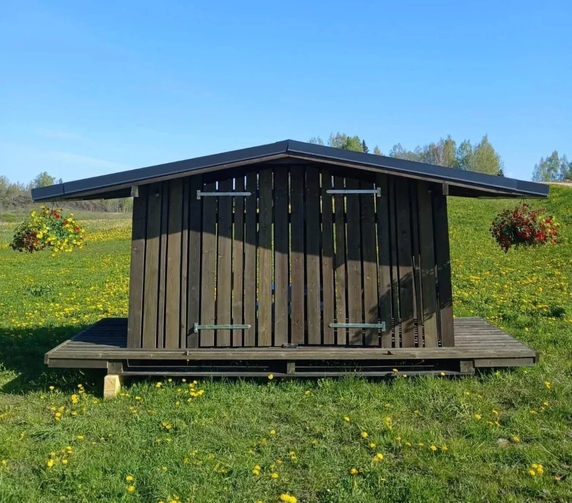 A small black wooden shed with a pitched roof, positioned on a grassy field with yellow flowers, and two hanging flower baskets on either side.