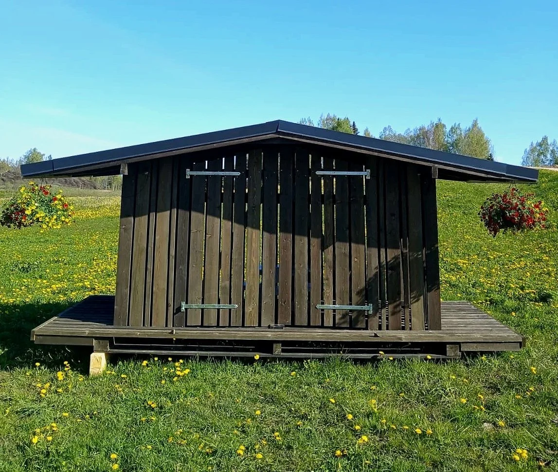 Small wooden shed with a sloped roof in a grassy field with yellow flowers, trees in the background, and hanging flower baskets on the sides.