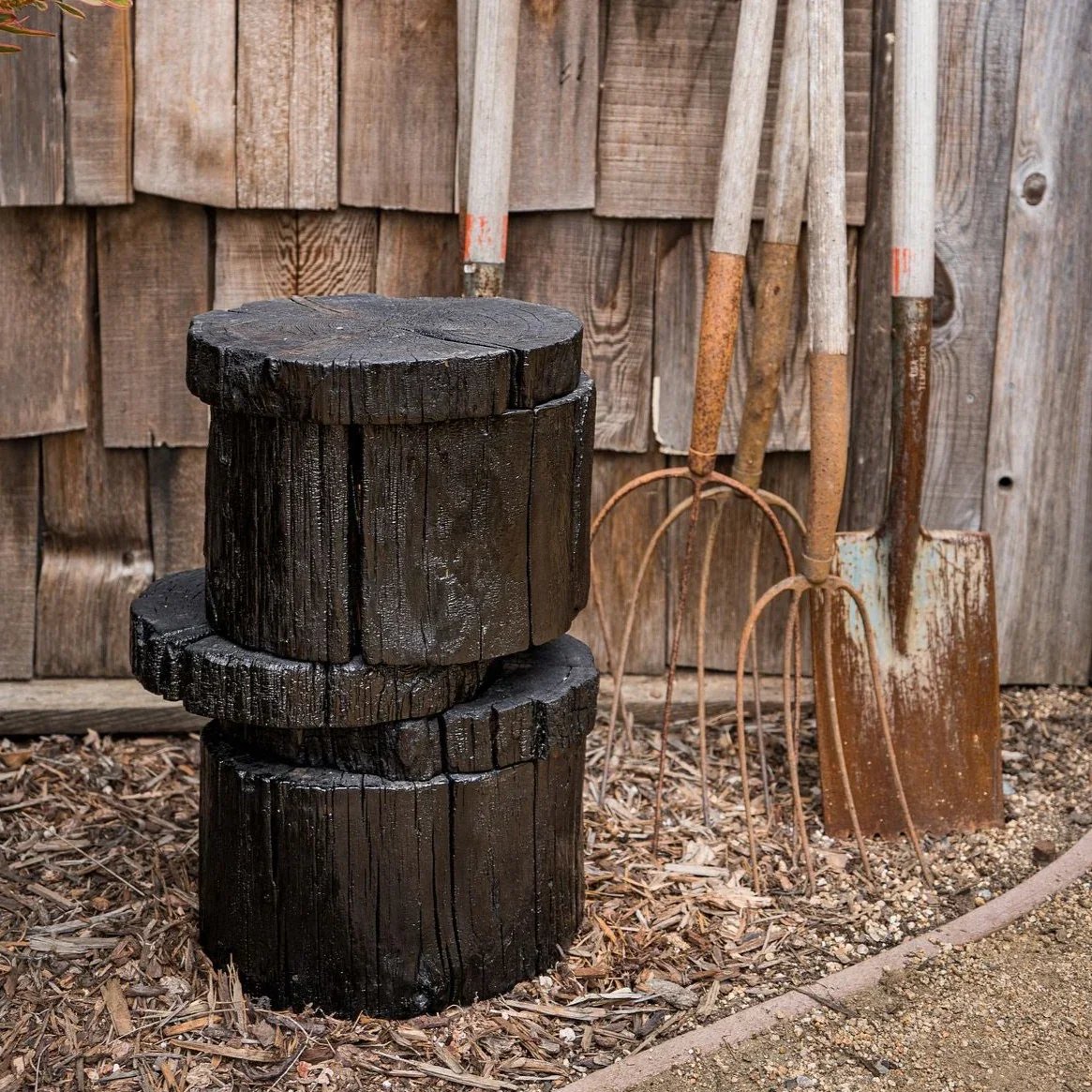 Set of three dark, burnt wood logs, topped with wood shavings, placed outdoors on the ground. Behind them, four rusty garden tools with wooden handles leaning against a weathered wooden fence.