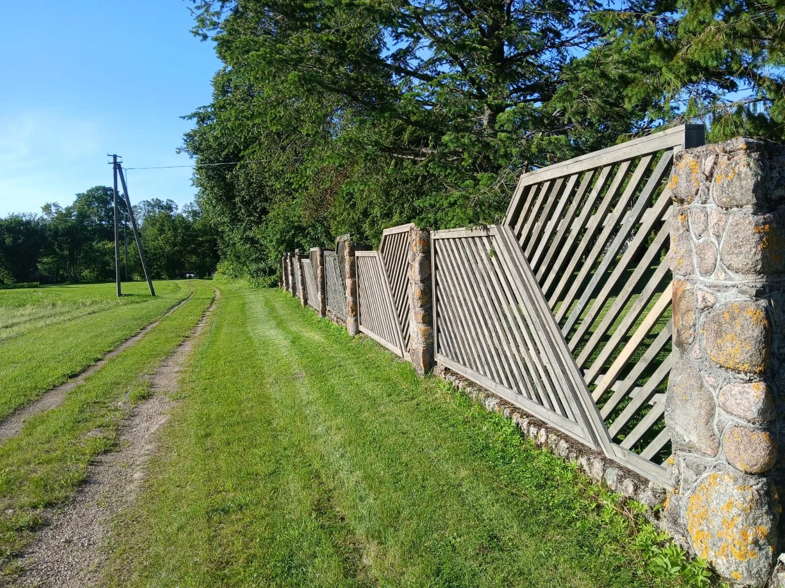 A wooden fence with stone posts runs along a grassy dirt path on a sunny day, with green trees and a utility pole in the background.