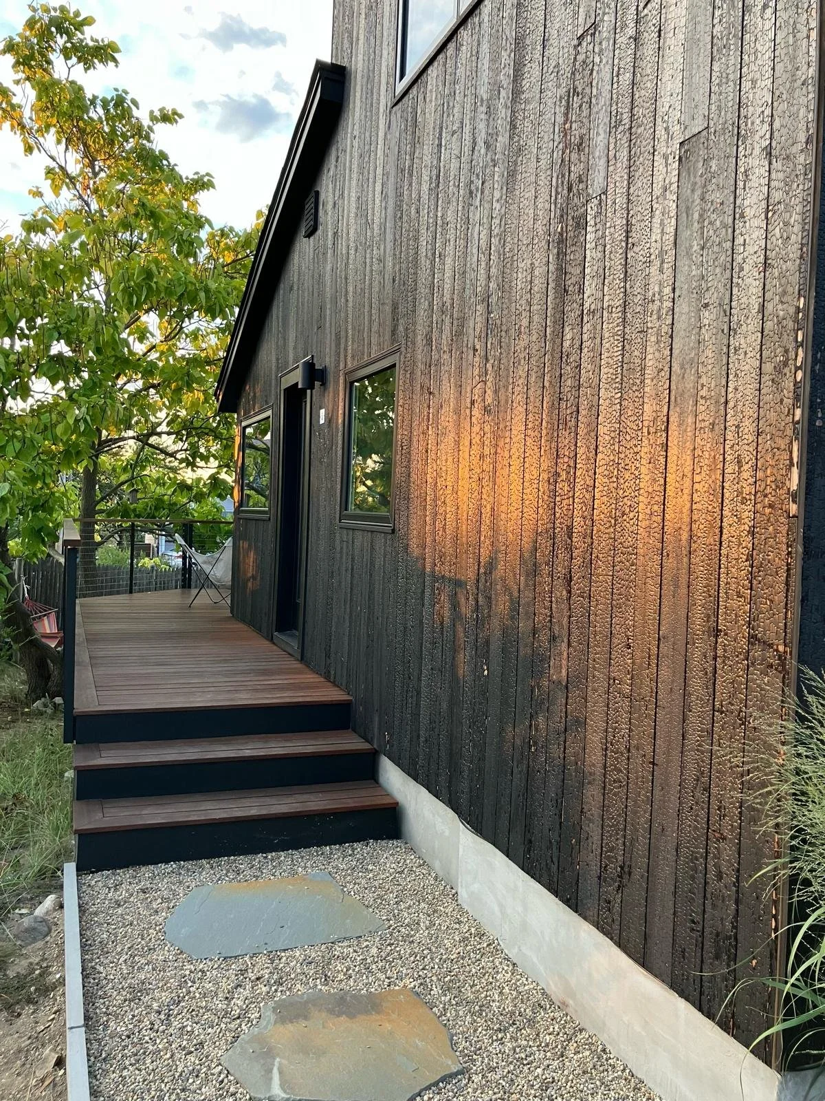 Side view of a modern house with charred wooden exterior, small steps leading to a door, and a backyard with trees and a hammock.