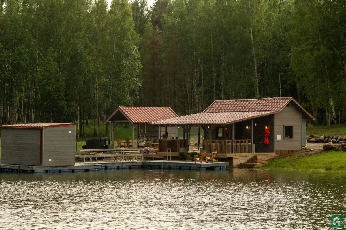 A house and a small cabin by a lake, with trees in the background, during daytime.