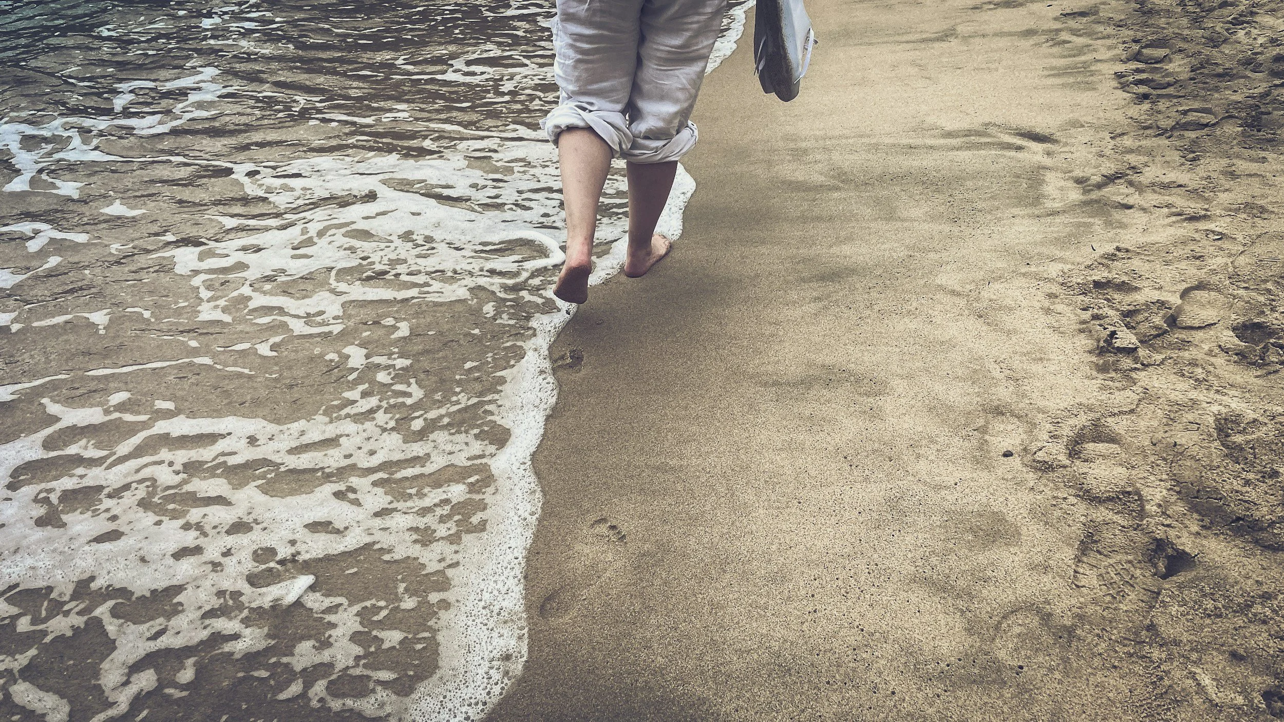 A person walking barefoot along the shoreline with waves gently washing over their feet on a sandy beach.