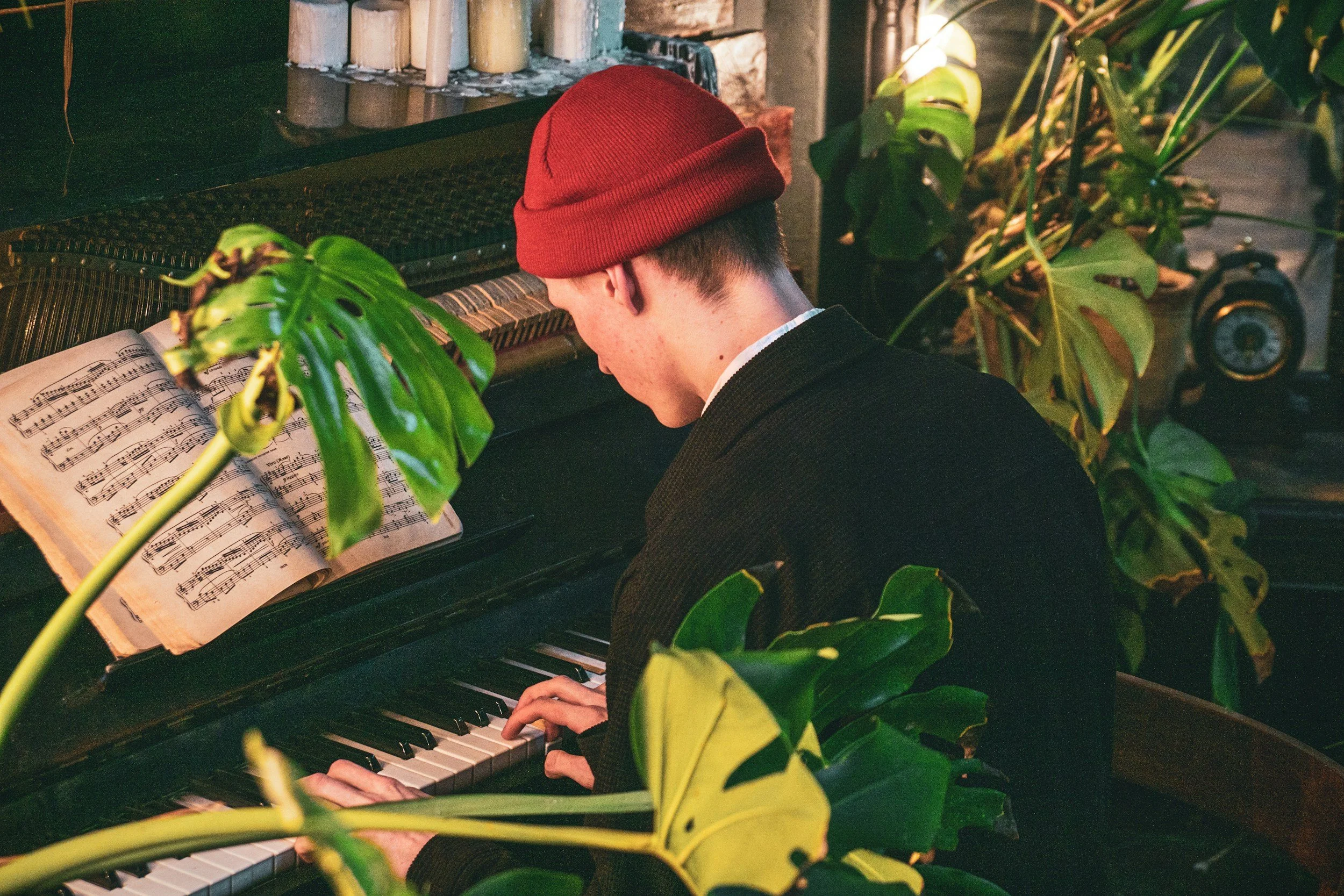 A man wearing a red beanie and black jacket playing a piano with sheet music open, surrounded by green leafy plants in a cozy indoor setting.