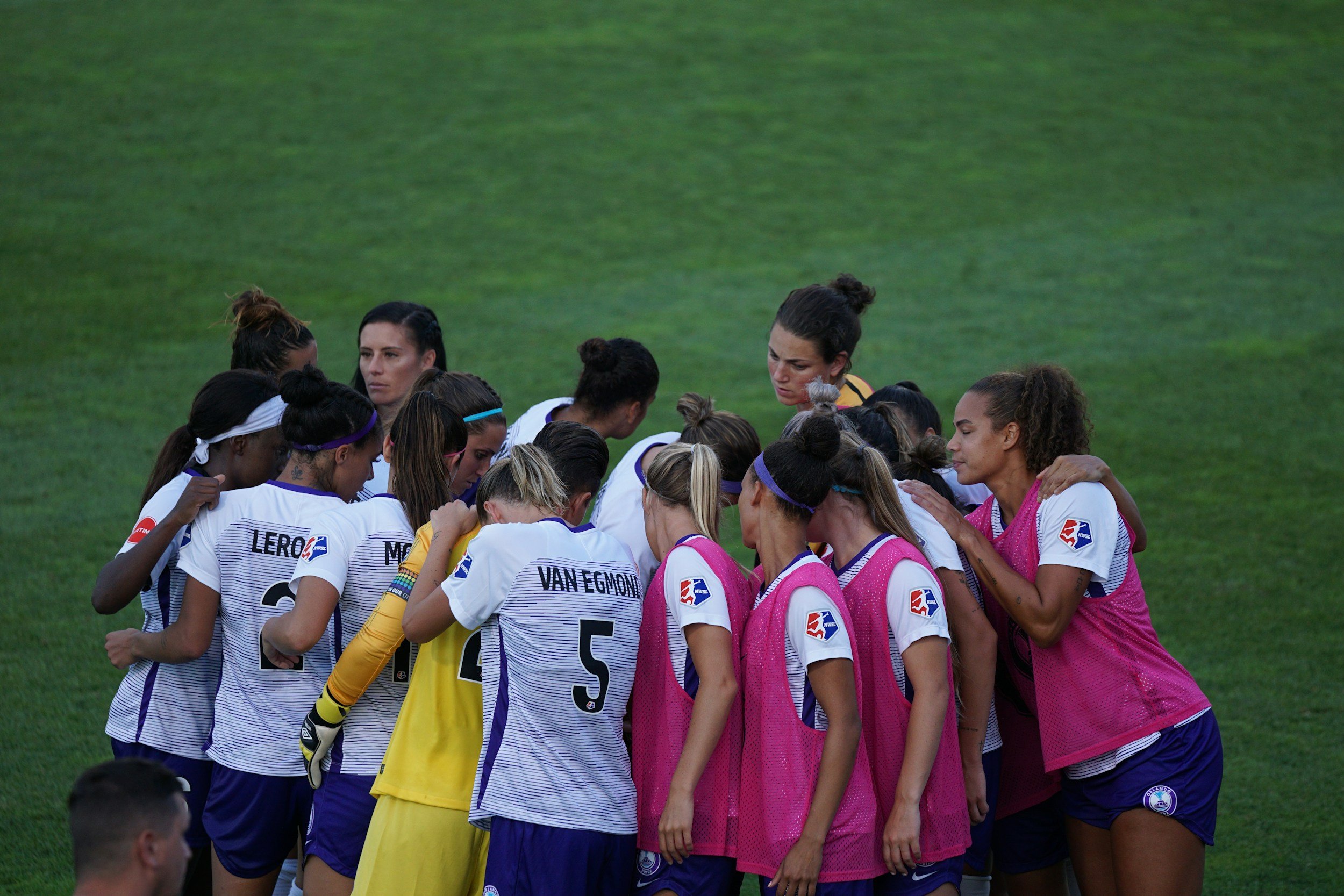 A group of female soccer players in white and pink jerseys huddled together on a green soccer field during a team meeting or pep talk.