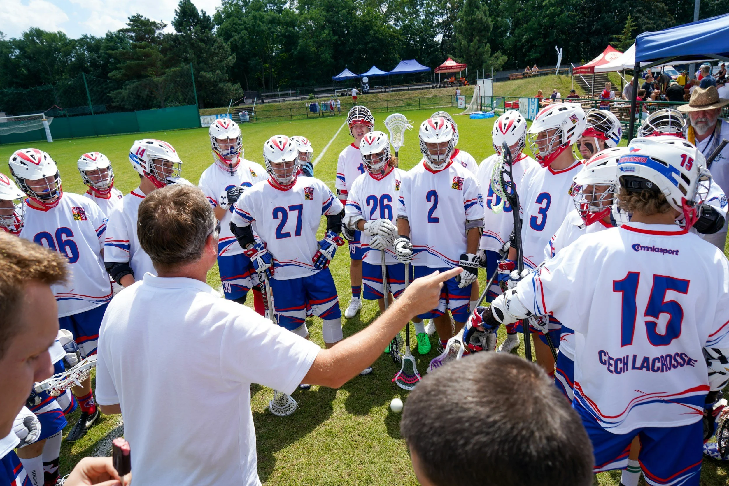 Lacrosse team in white jerseys with blue and red accents in a huddle on a grassy field, listening to coach during daytime.