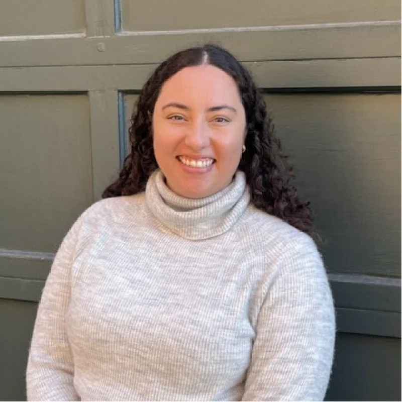 A woman with curly dark hair smiling, wearing a cream-colored turtleneck sweater, against a green paneled wall.