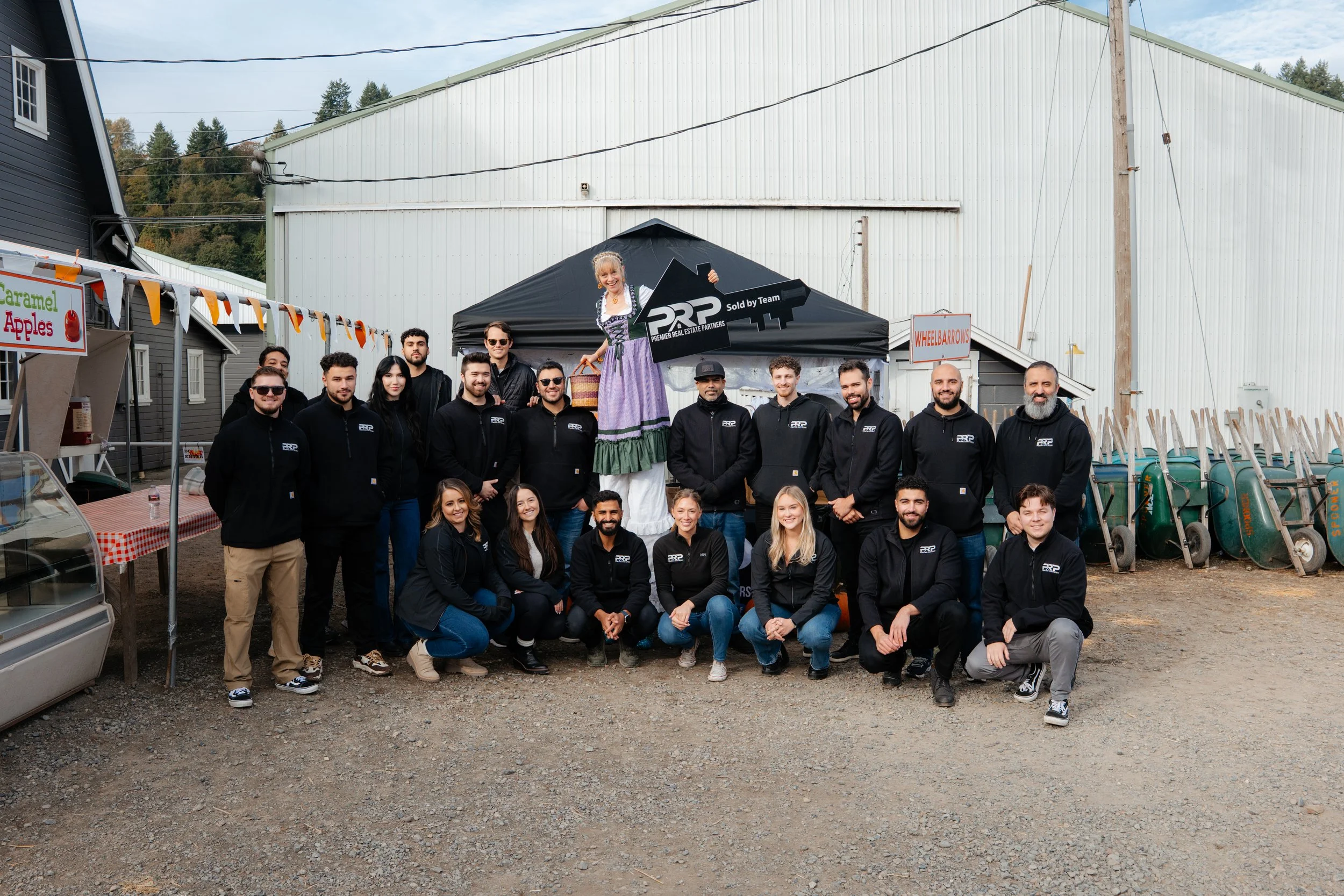A group of fifteen people posing outdoors in front of a black tent with a figure dressed as a traditional as Saint Lucia in a purple dress and a crown of candles. The group is wearing black hoodies with a white logo. There are signs for caramel apples and wheelbarrows, and a small tent or booth to the left, with trees in the background.