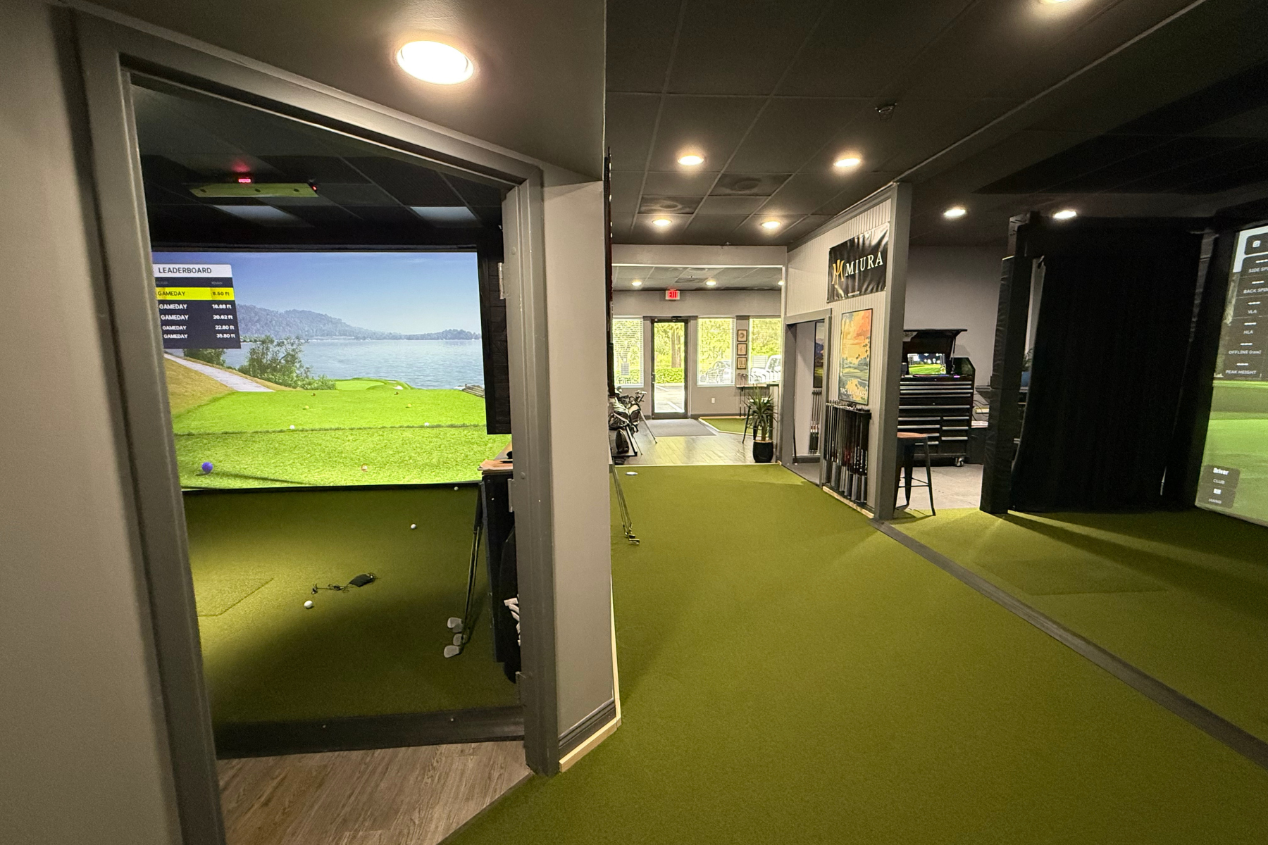 Indoor golf simulation room with a large screen displaying a golf course view, golf clubs, and equipment on the floor. Green turf flooring, and the room is well-lit with ceiling lights.