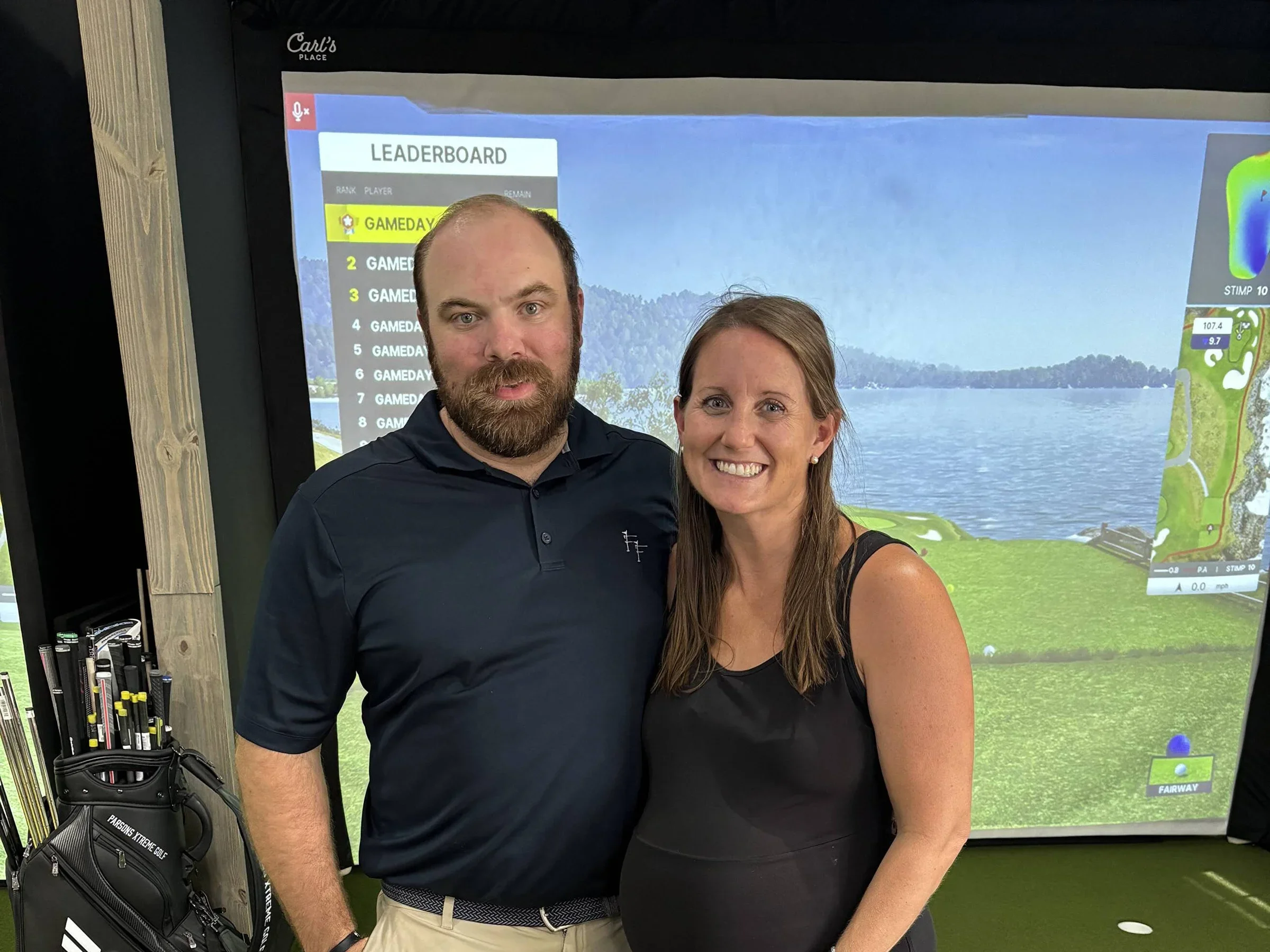 A man and woman standing together, smiling indoors in front of a golf simulator screen displaying a scenic water and land view, with a golf bag and clubs nearby.