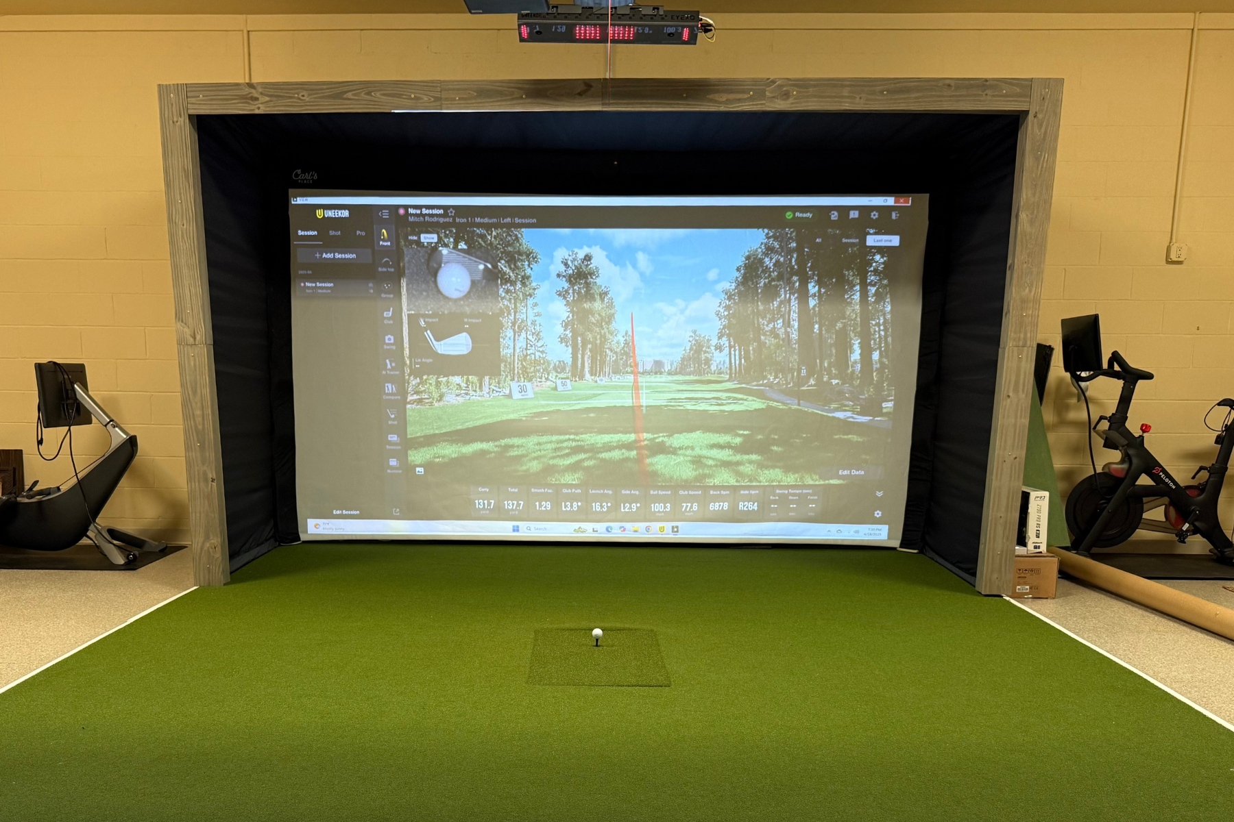 Indoor golf simulator with orange tee and green artificial turf, displaying a virtual golf course entrance with trees and sky.