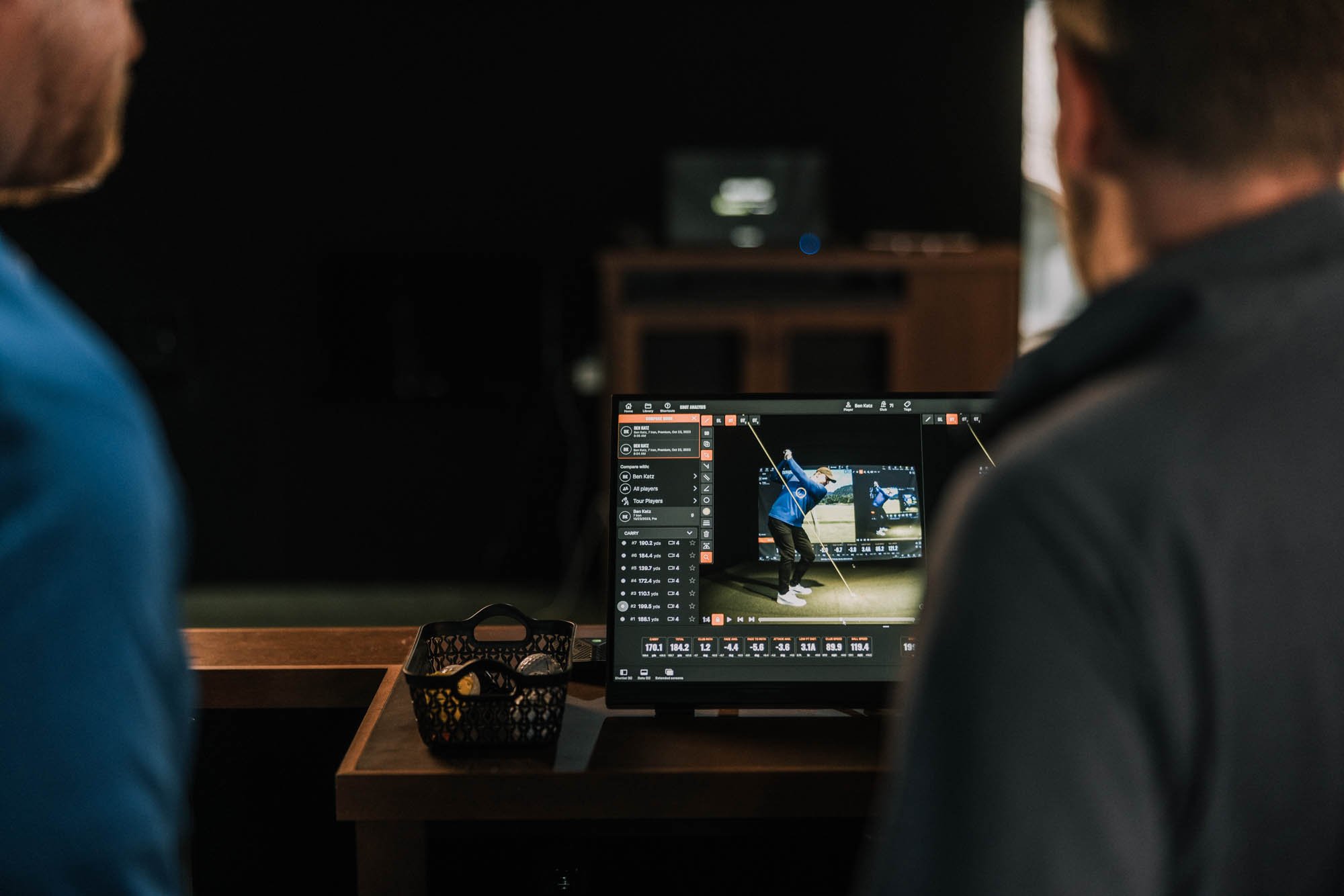 Two people observing a computer screen showing a golf swing analysis with video clips and data charts in a dimly lit room.
