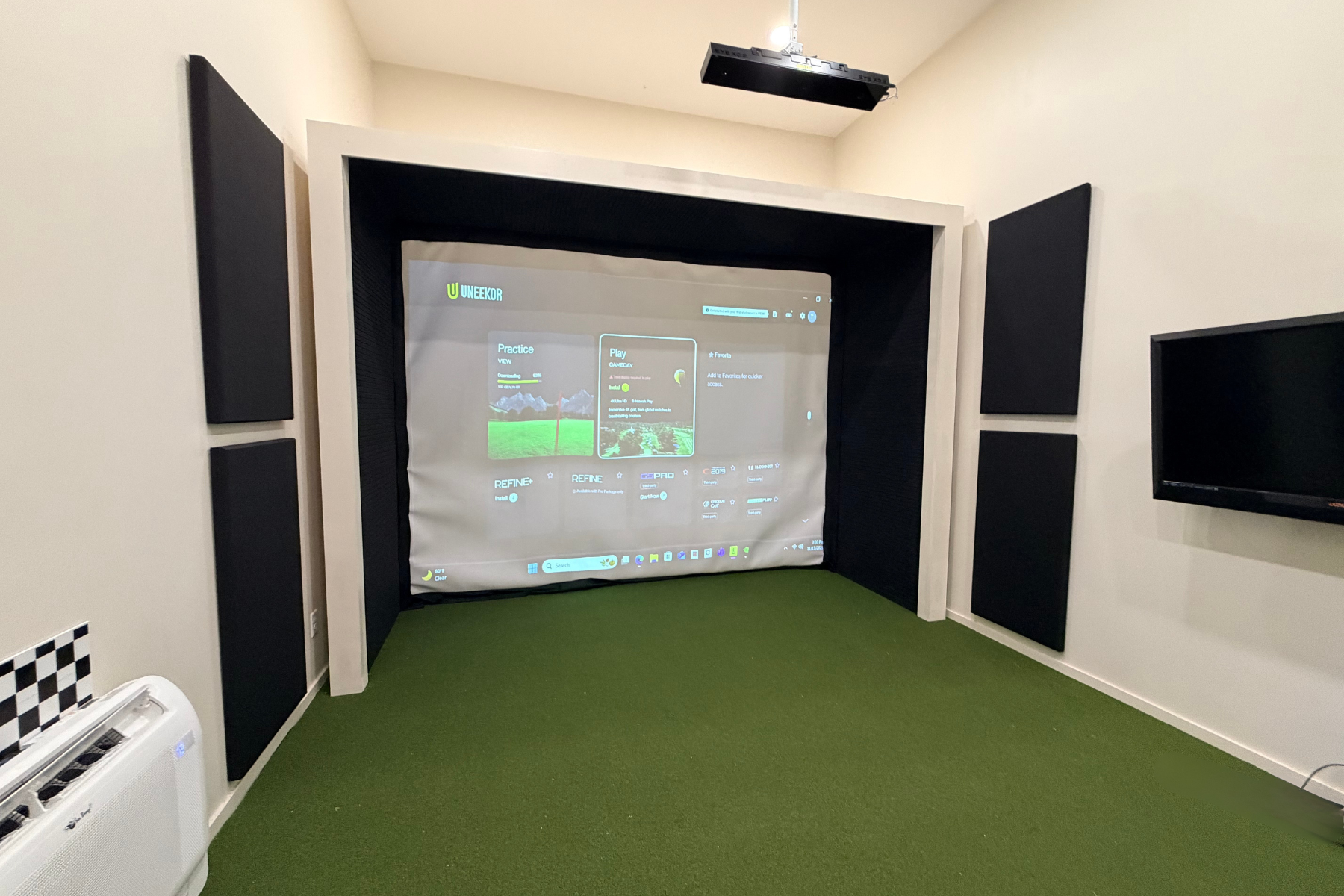 Indoor golf simulator room with a large projection screen, black acoustic panels on the walls, a green carpet, and a flat-screen TV on the wall.
