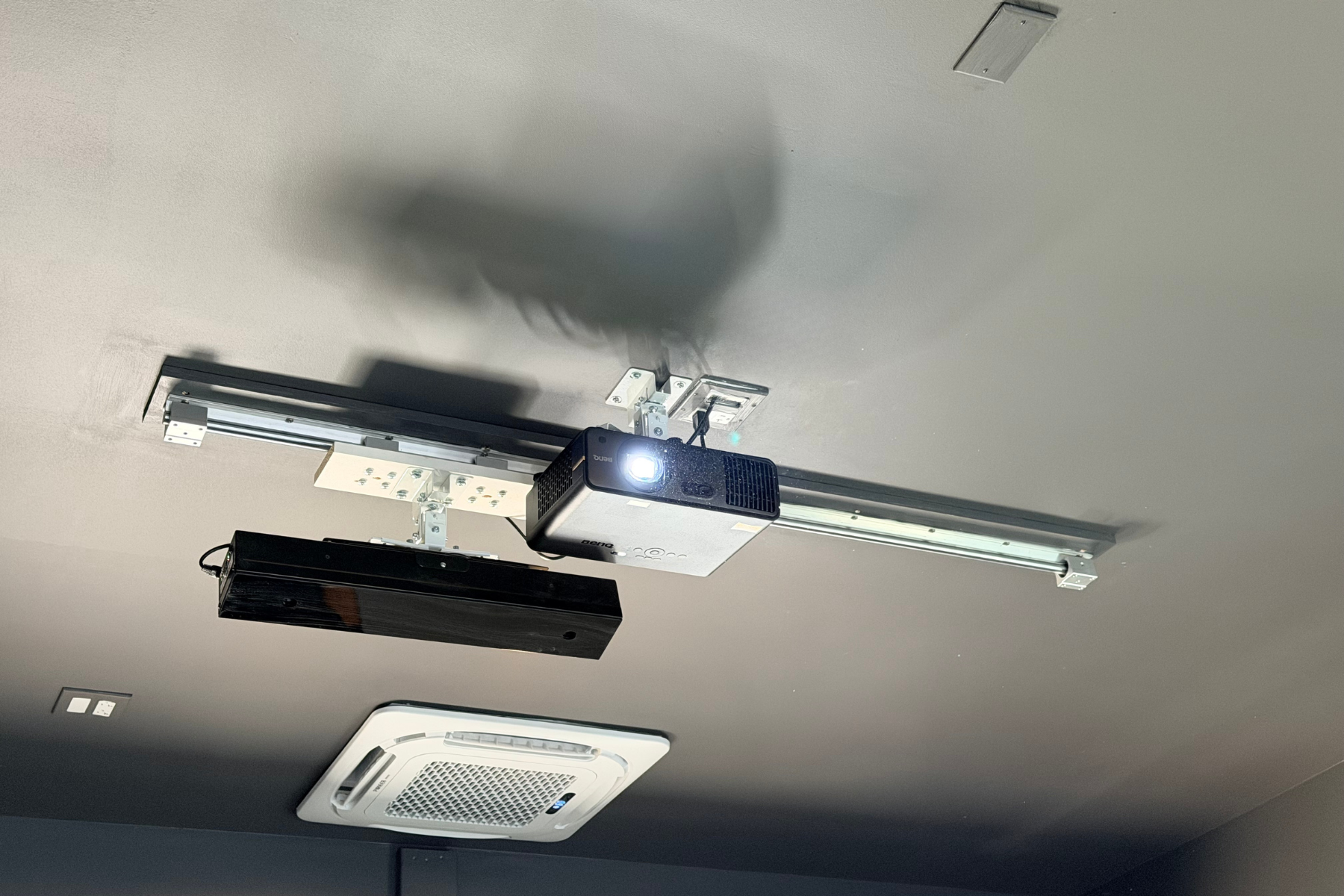 Ceiling-mounted projector with a bright lens and sliding golf launch monitor, and a white air conditioning vent on the ceiling.