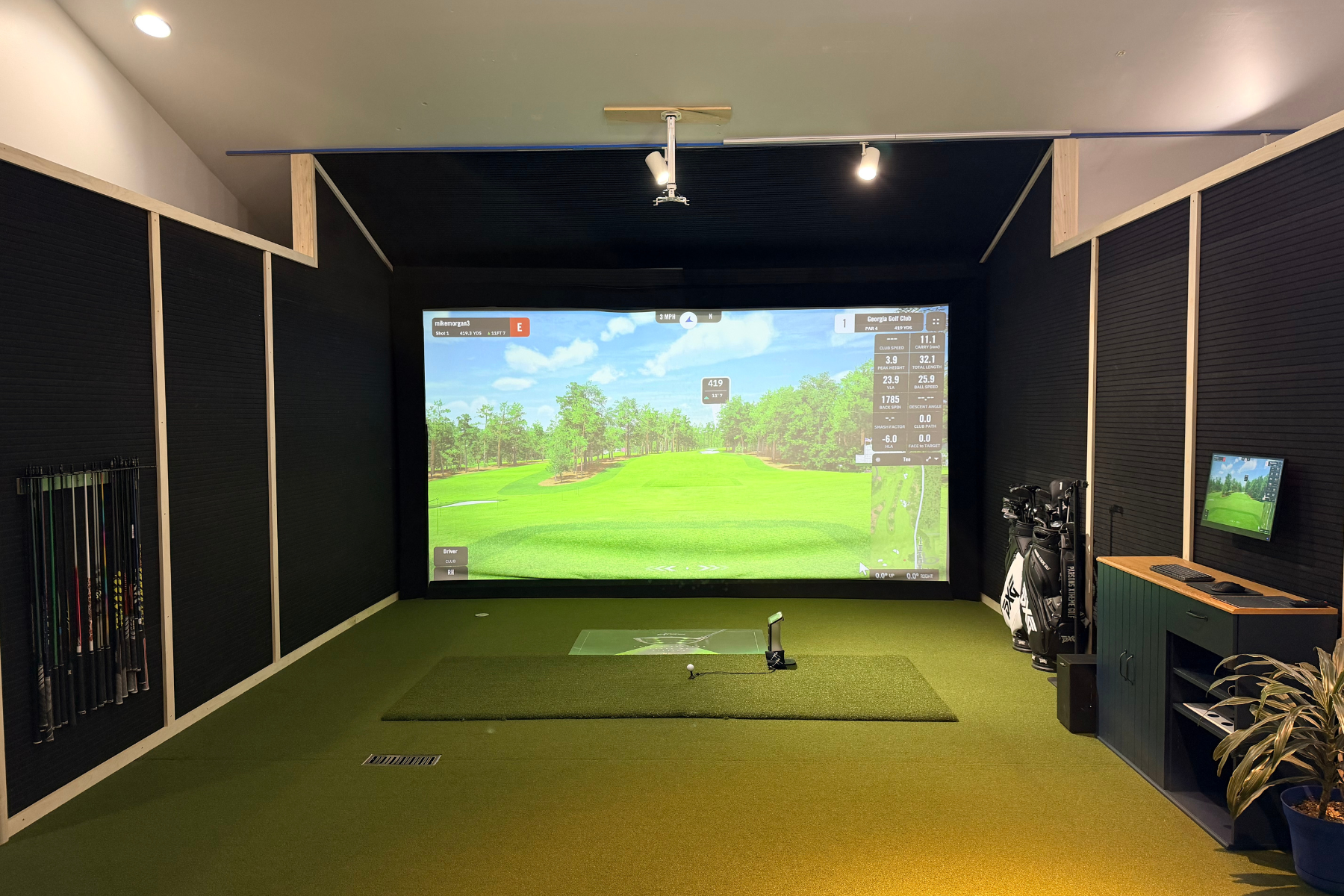 Indoor golf simulator room with screen displaying a virtual golf course, golf clubs in a stand, and a small monitor on a desk.