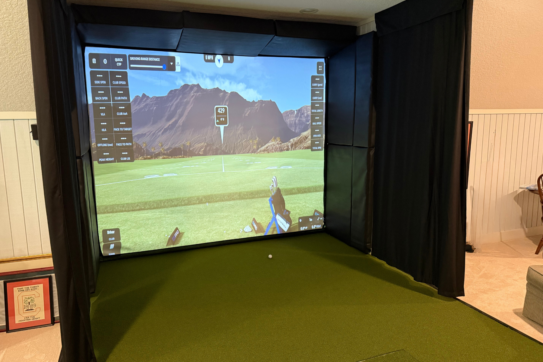 Indoor golf simulator setup with a large screen displaying a virtual golf course, enclosed by black curtains, with a green turf floor and a small golf ball on the floor.