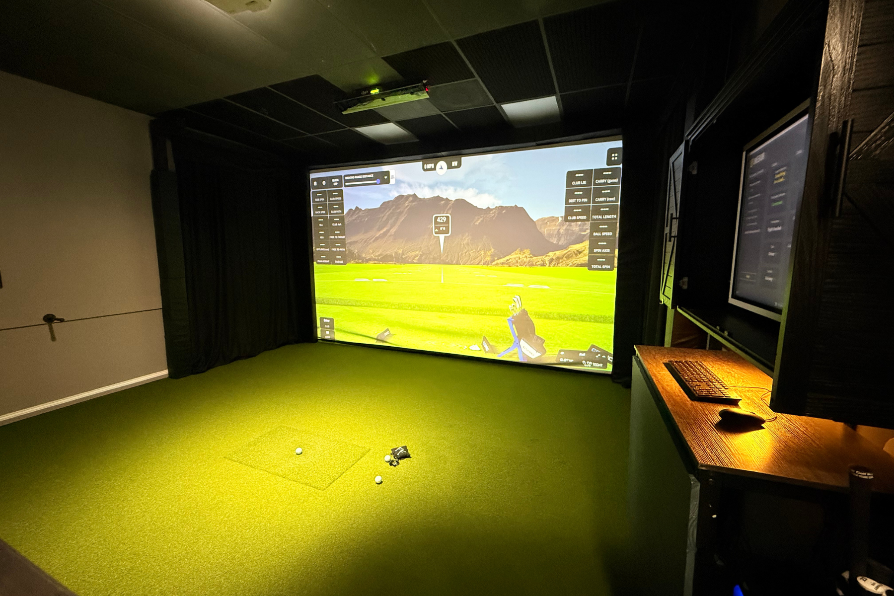Indoor golf simulator room with a large screen displaying a virtual golf course, green carpeted floor, a golf ball, and a golf club on the floor. There is a desk with a computer and monitor on the right side.