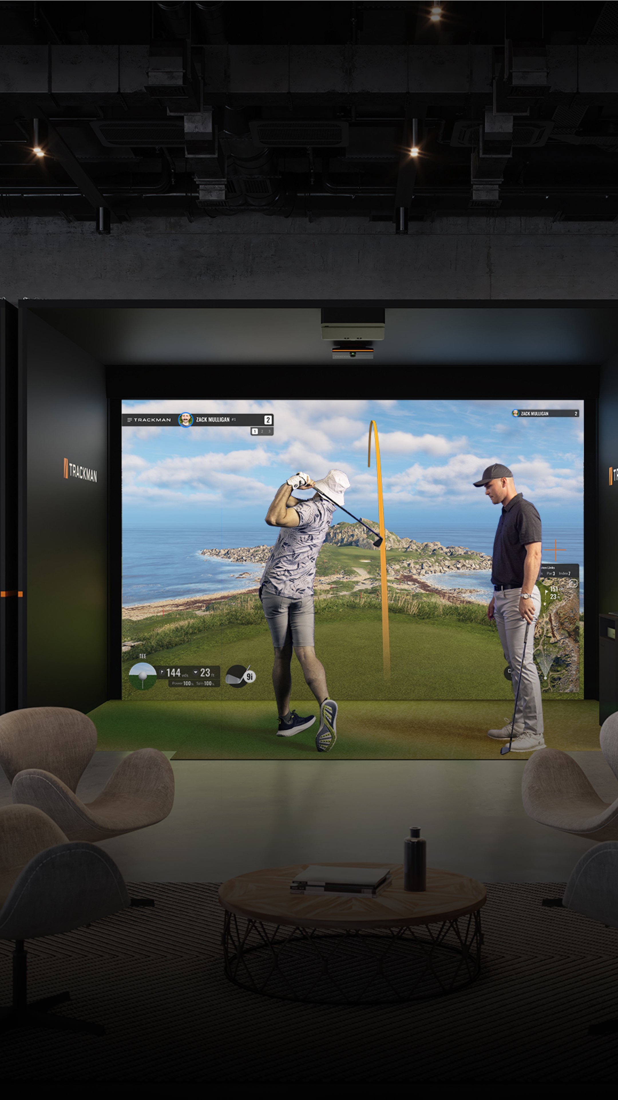 Indoor golf simulation room with a large screen displaying a golf course, two men playing golf, one swinging a club and the other watching.