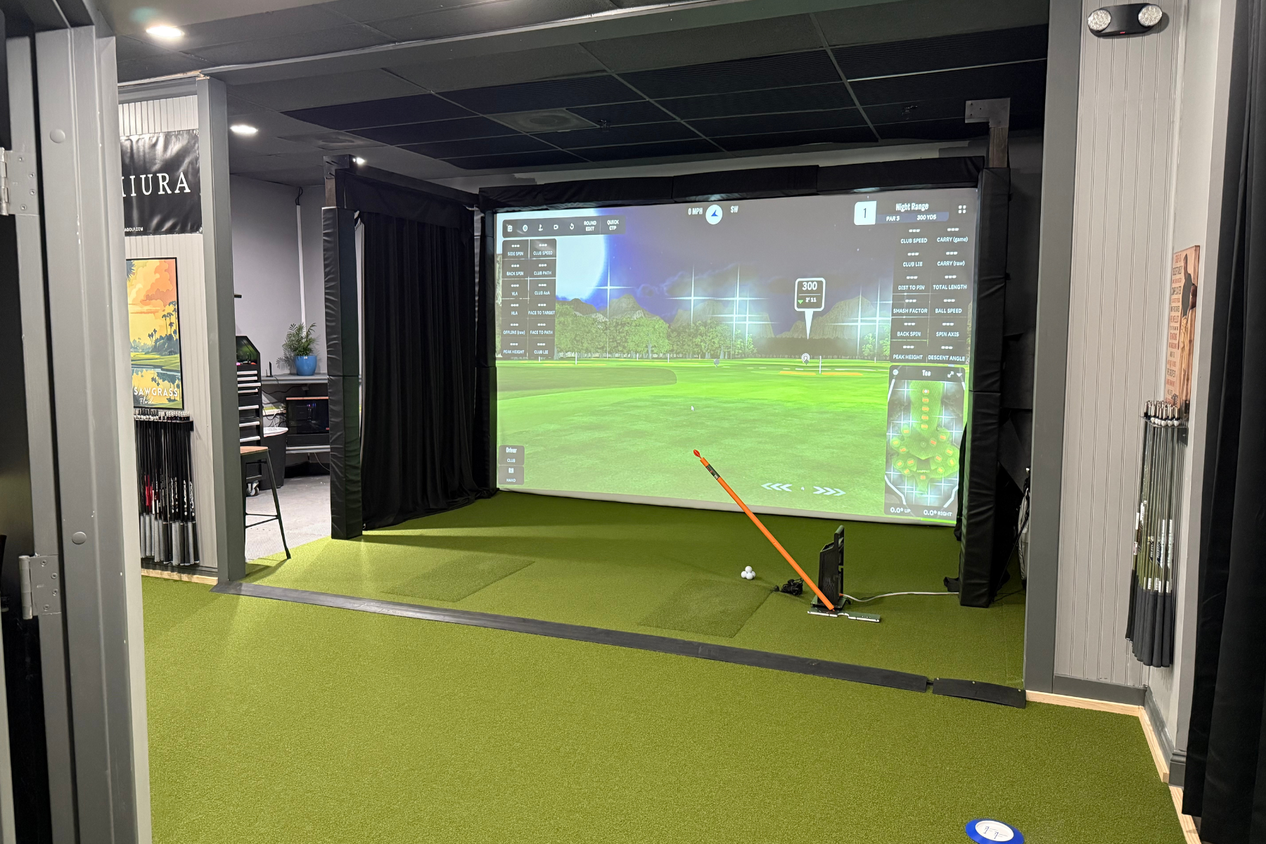 Indoor golf simulation room with a large screen displaying a virtual golf course. There is a golf club and two golf balls on the green mat in front of the screen, and various golf clubs are stored to the left. The setup includes a projector, a small 