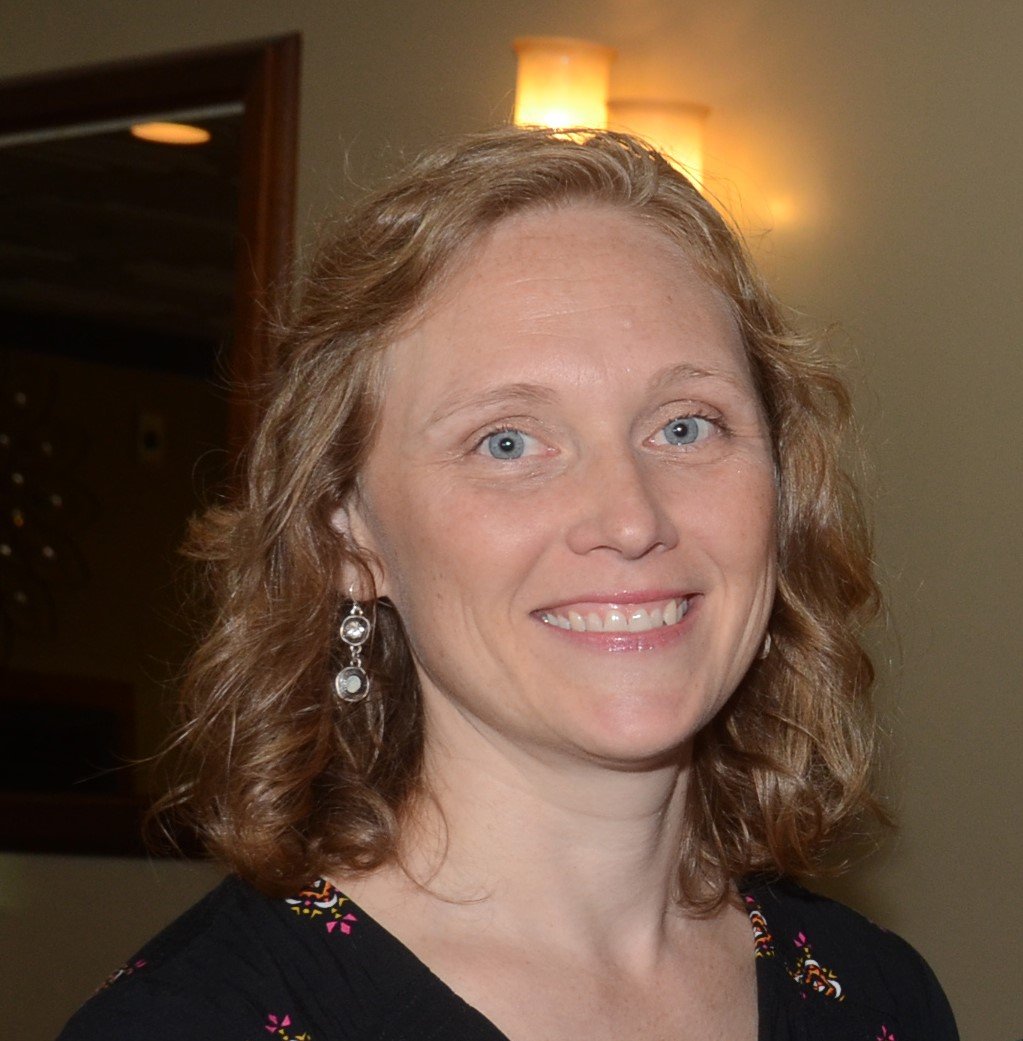 A woman with curly red hair, blue eyes, and earrings, smiling at the camera in a warmly lit indoor setting.