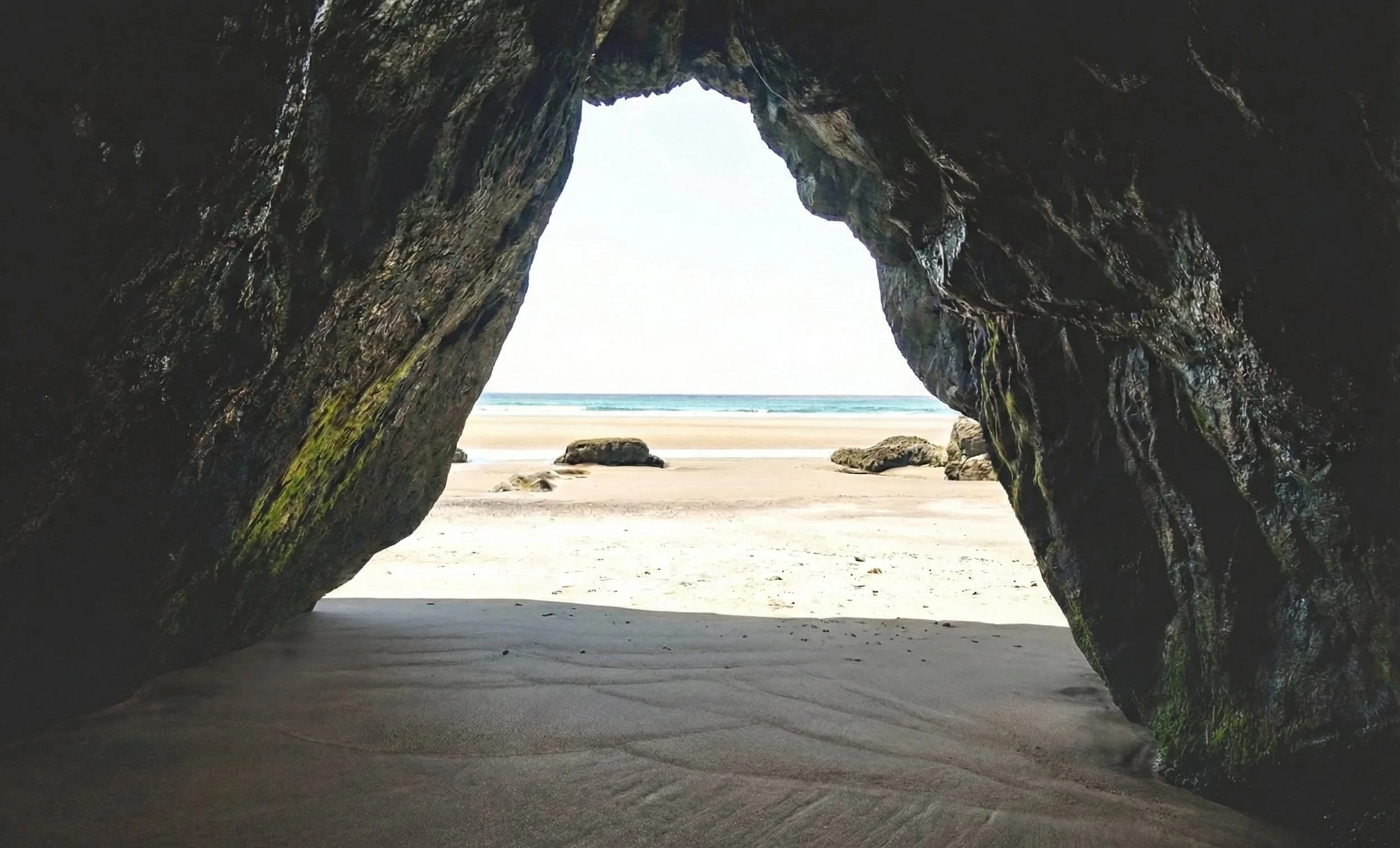 A view from inside a dark cave looking out onto a pristine, sunlit beach and ocean horizon, representing the limitless potential of Elite Redox telehealth services.