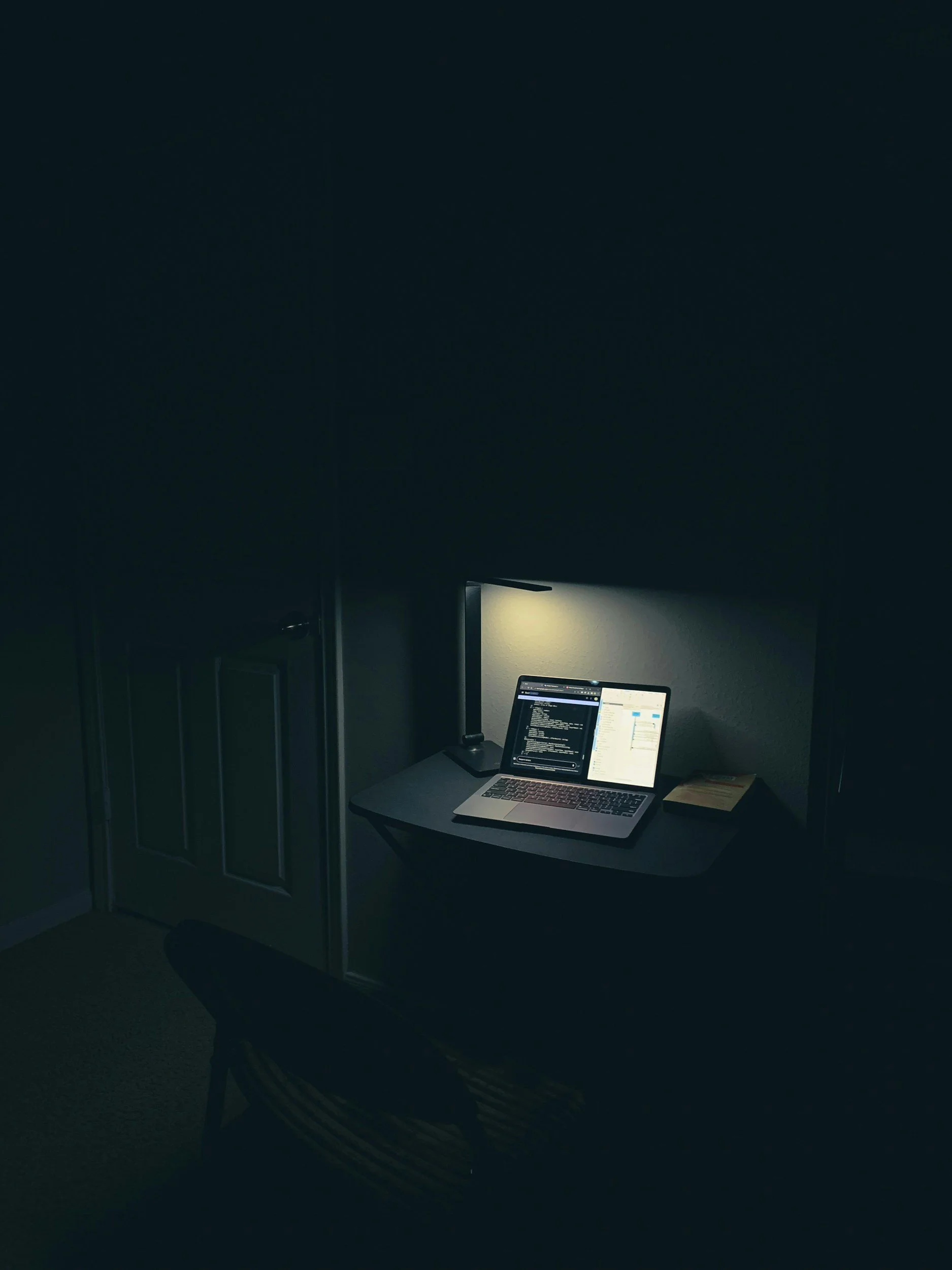 A laptop on a desk in a dark room, illuminated by a small desk lamp, with code displayed on the laptop screen.
