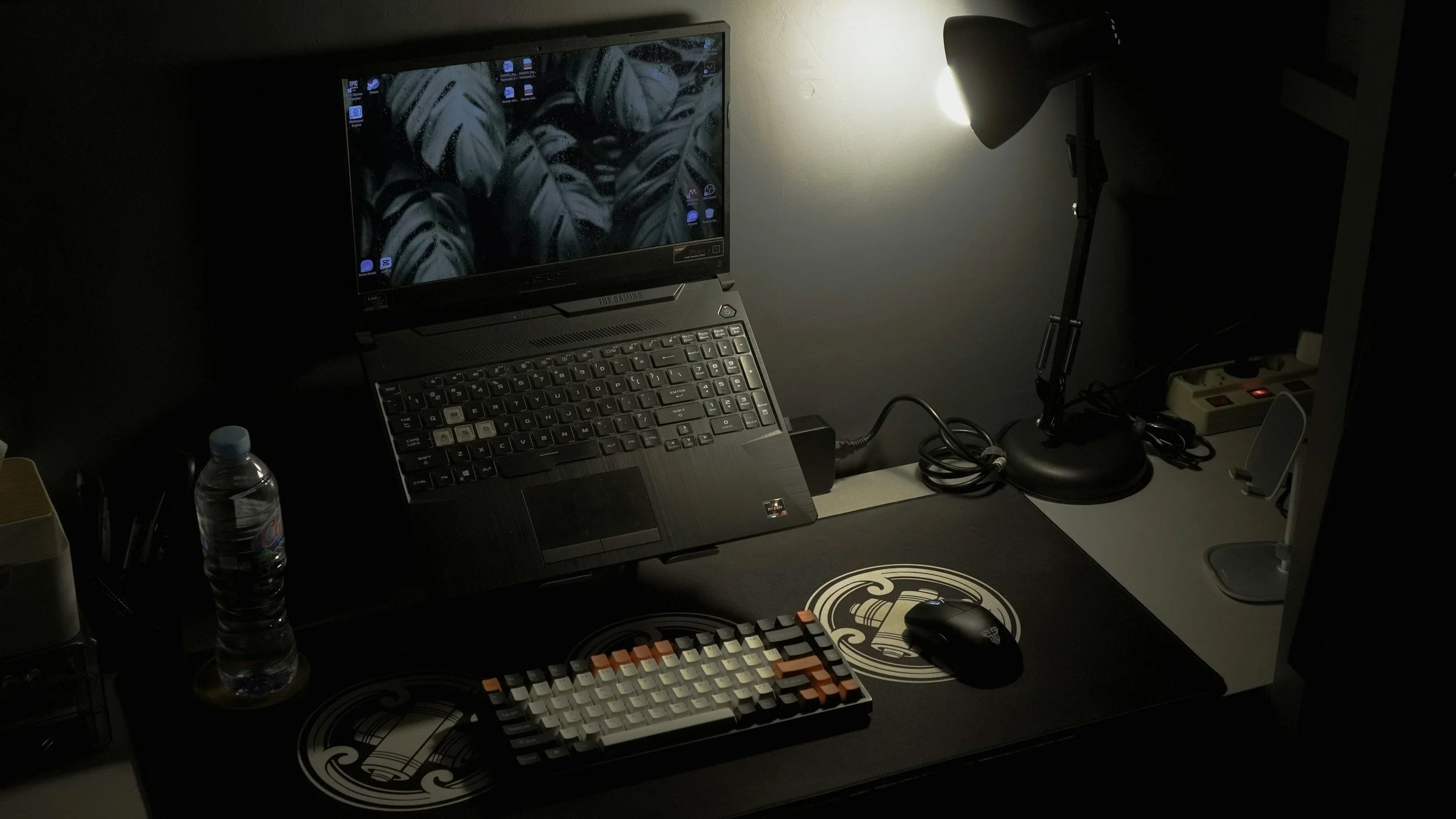 A dark workspace with a computer monitor displaying a leaf-themed wallpaper, a laptop, a keyboard with orange and cream keys, a mouse, a desk lamp, a water bottle, and various accessories on a black desk mat with an ornate design.