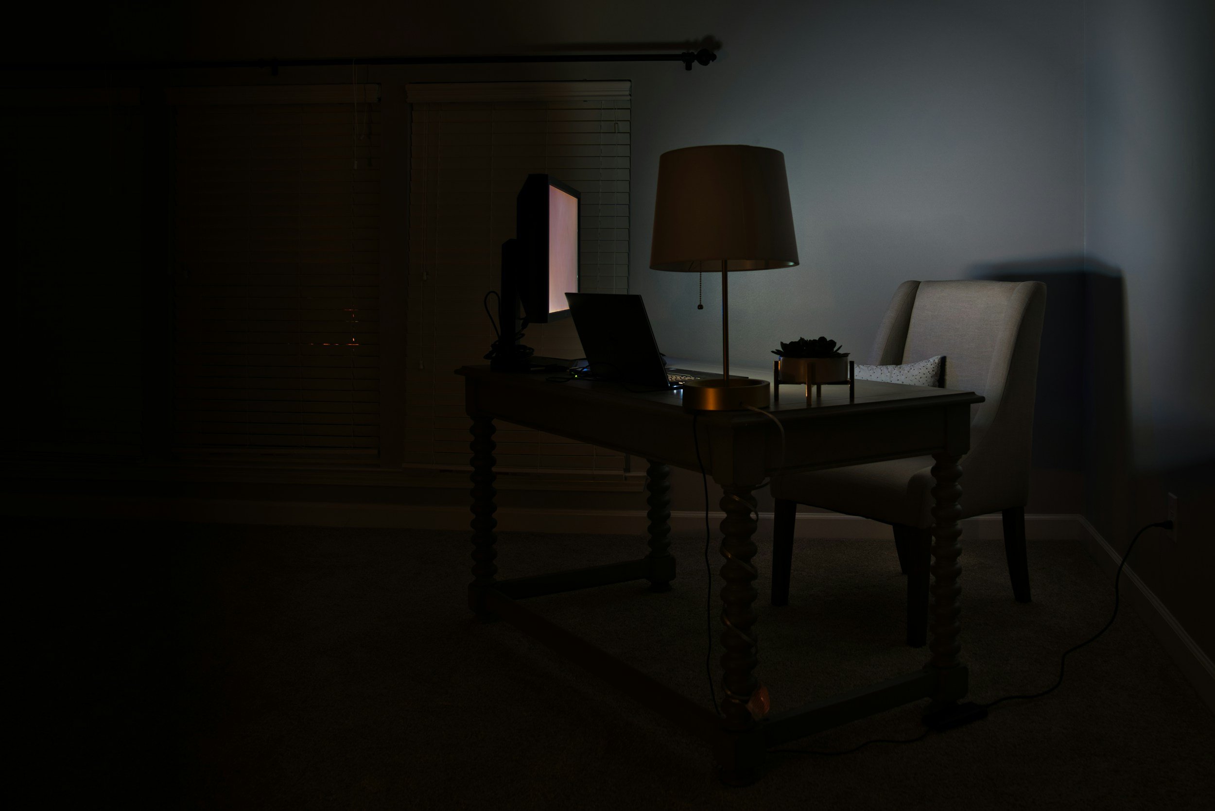 Dimly lit home office with a wooden desk, a chair, a desk lamp, a laptop, a computer monitor, and some decorative items.