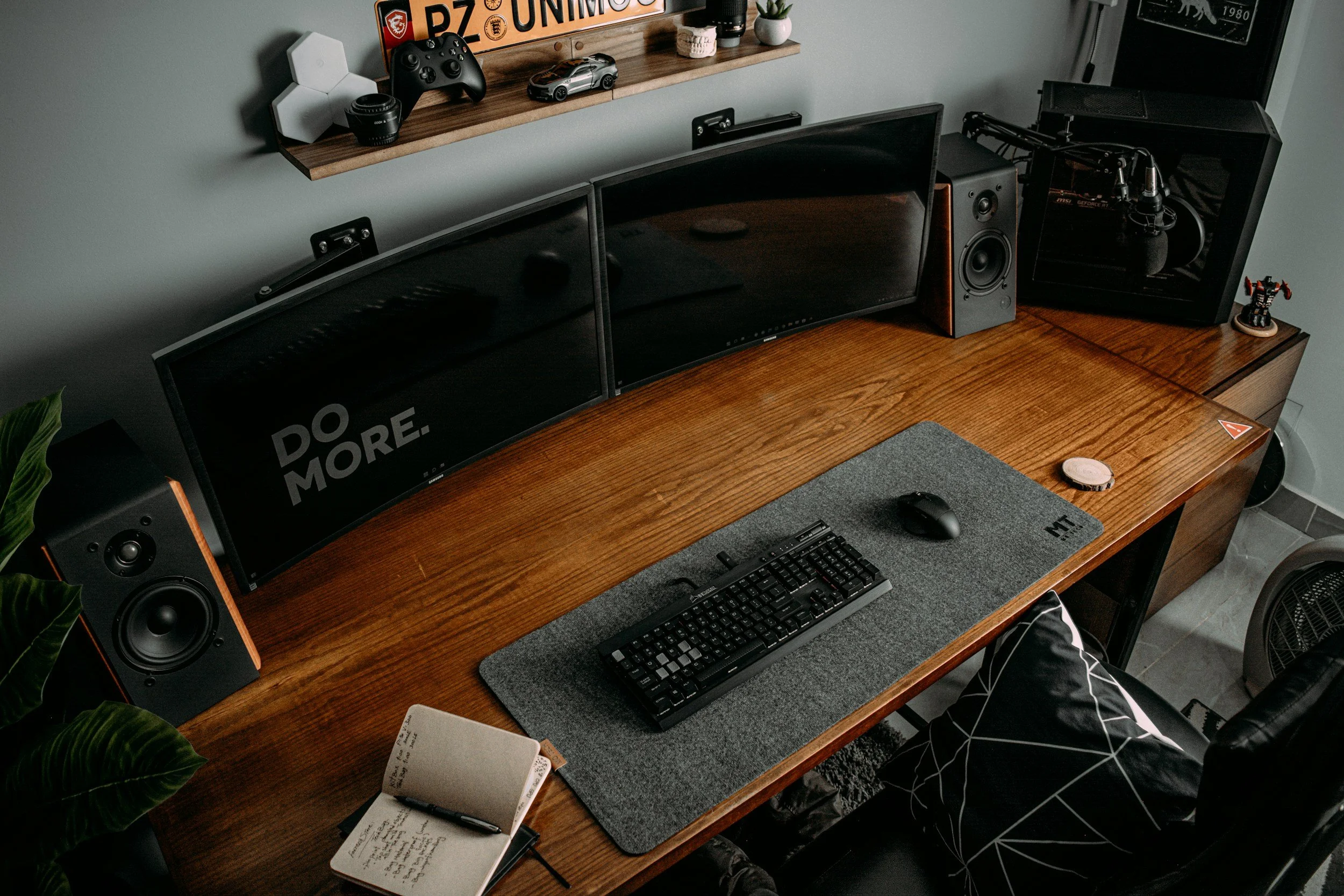 A wooden desk with three curved computer monitors, a keyboard, and a mouse on a gray desk mat. There are speakers on either side of the monitors, a notebook with writing and a pen on the left, and various decor items including collectible figures and a small potted plant. A shelf above holds models, gaming controllers, and framed art.