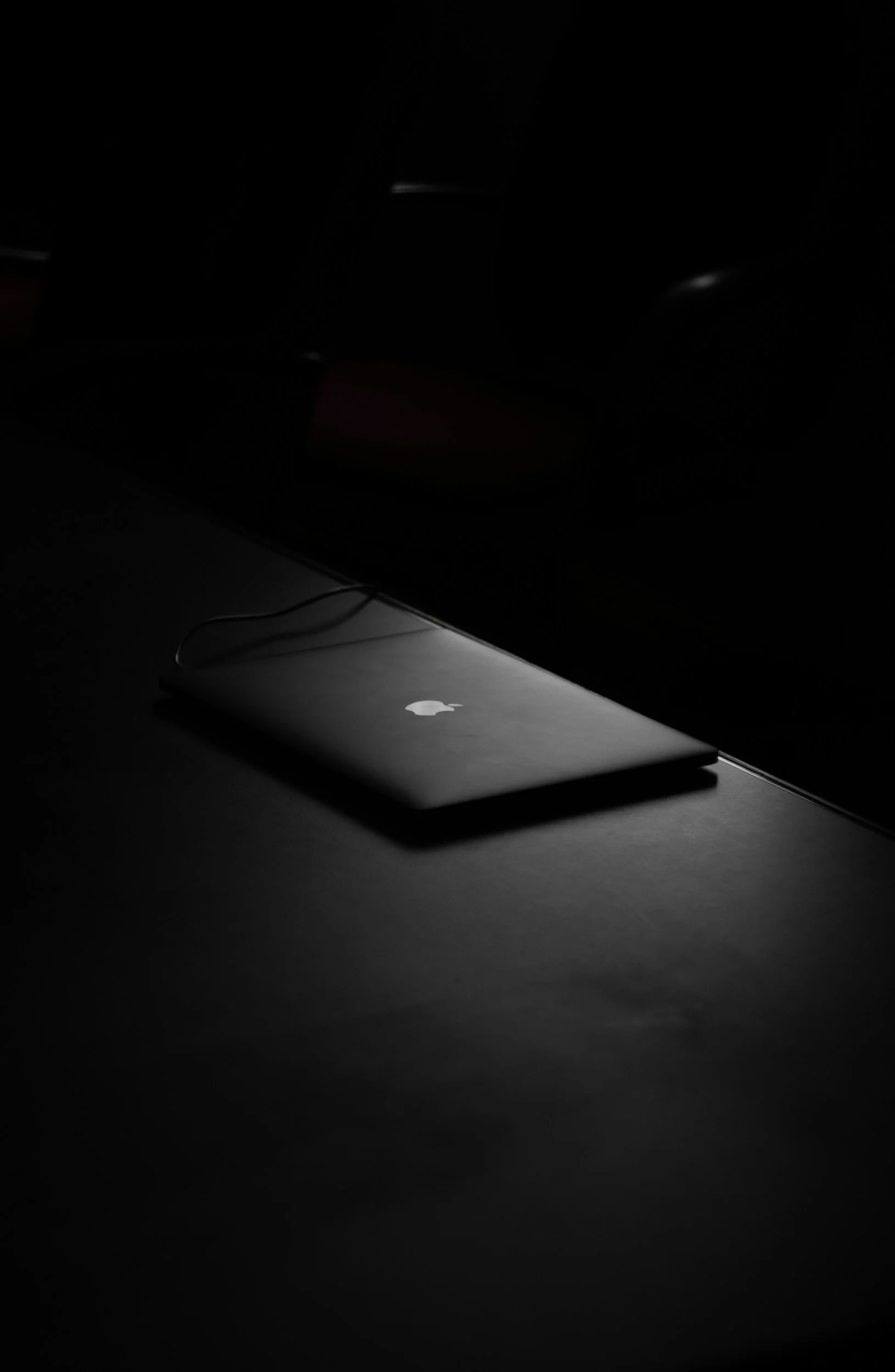 A closed MacBook laptop with an Apple logo on the lid, resting on a dark surface, illuminated by minimal lighting.