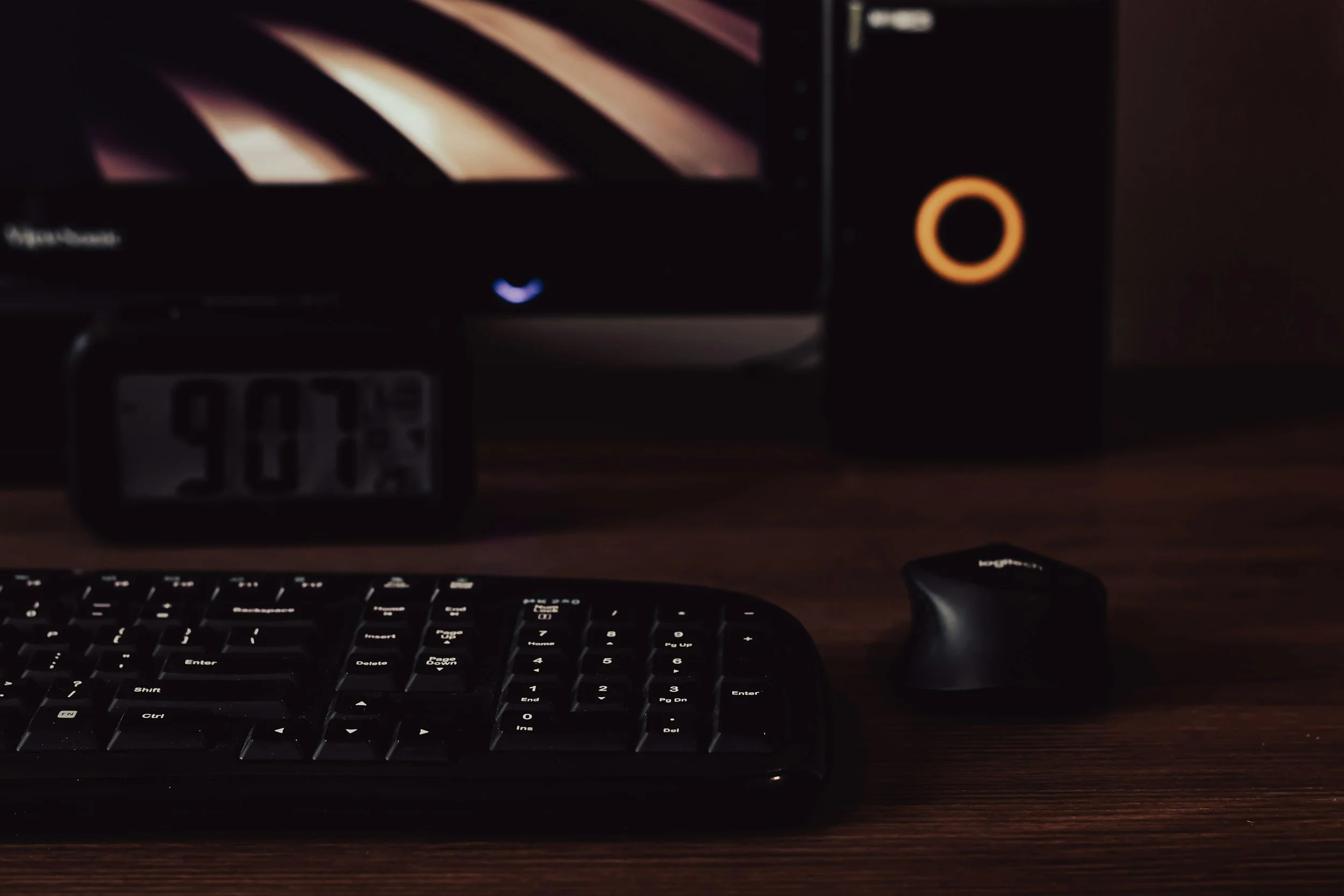 A computer keyboard, a wireless mouse, and a digital clock on a wooden desk, with a monitor and a speaker in the background.