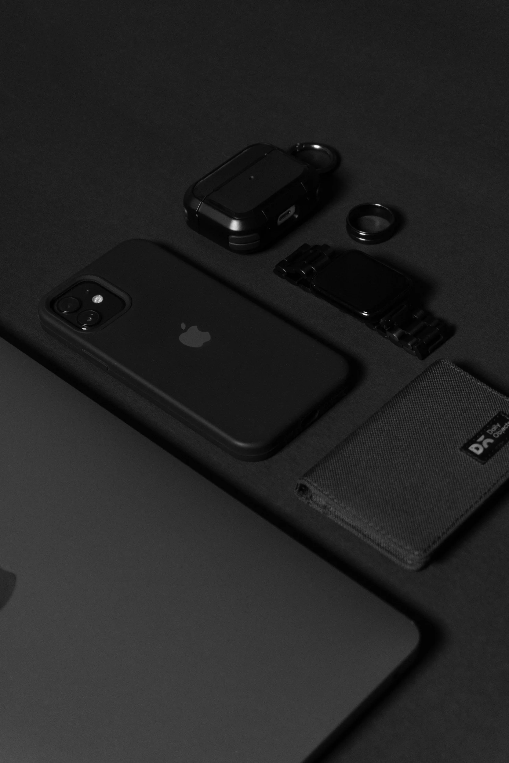 Black iPhone, wireless earbuds, smartwatch, ring, wallet, and laptop on a dark surface.