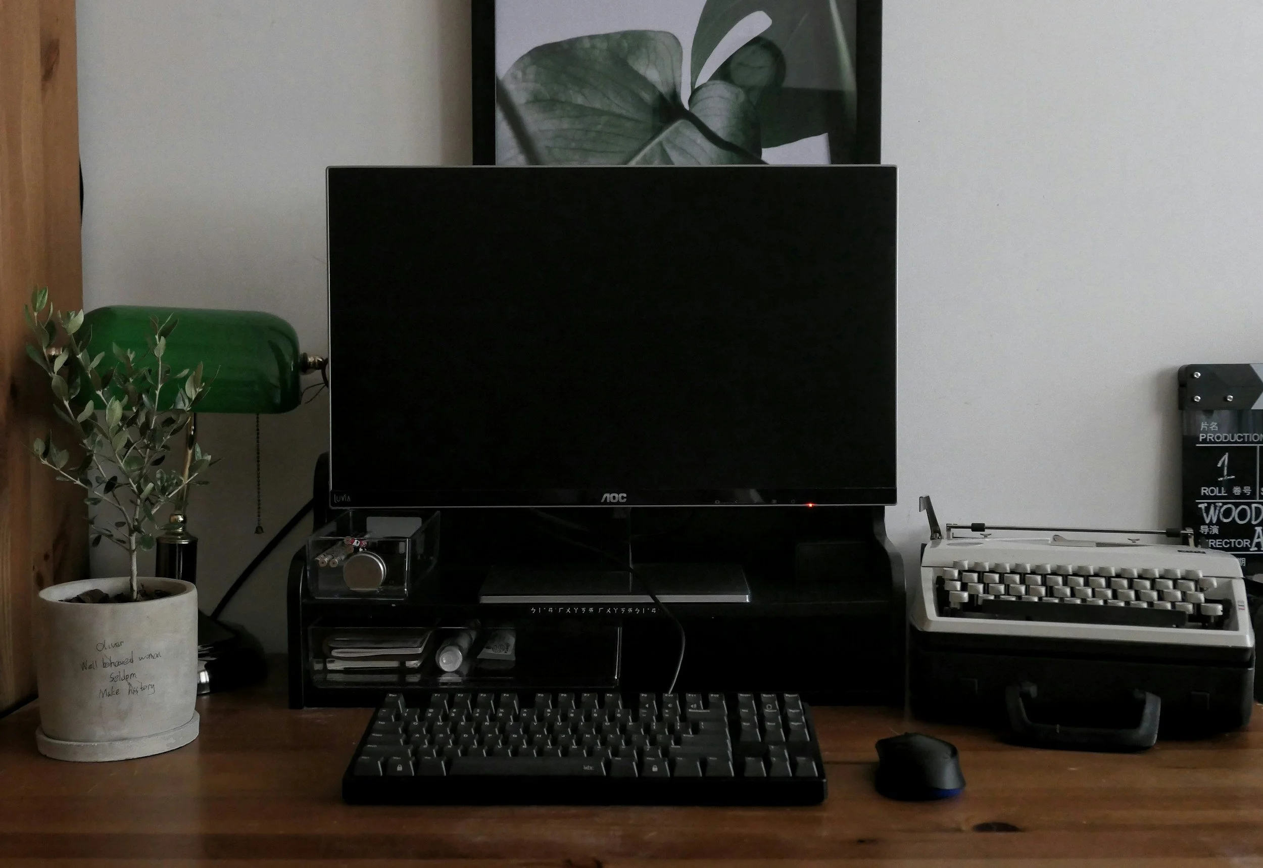 A home workspace with a wooden desk holding a computer monitor, keyboard, mouse, vintage typewriter, small potted plant, green desk lamp, and framed botanical art on the wall.