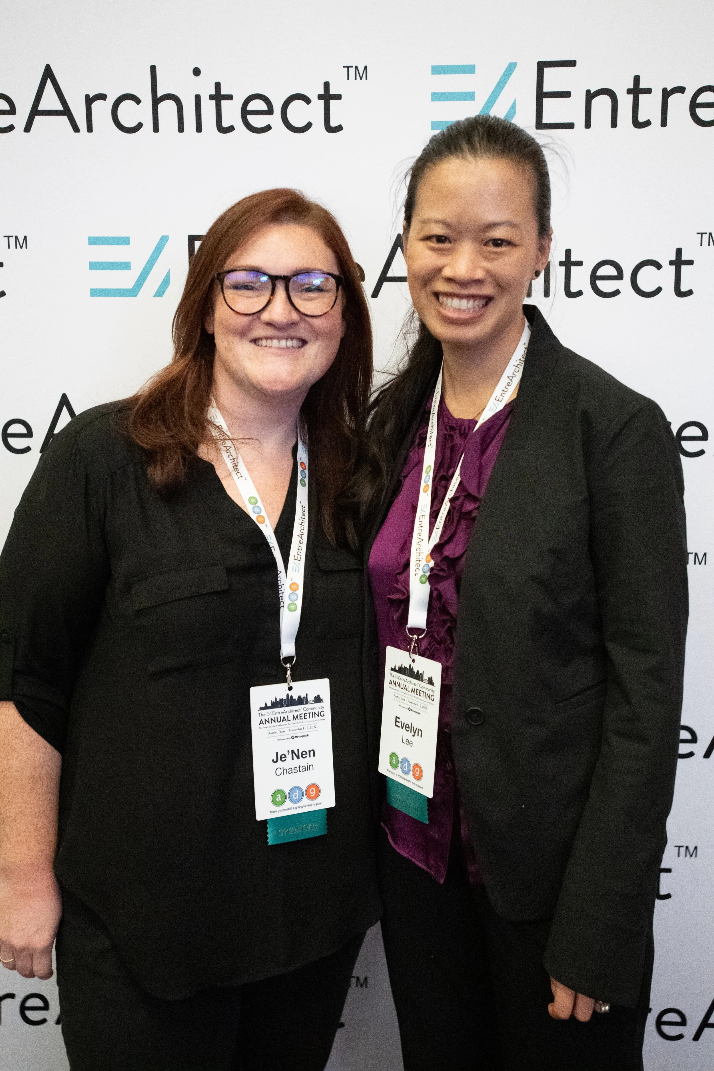 Two women smiling at a conference, wearing name tags and standing in front of a branded backdrop.