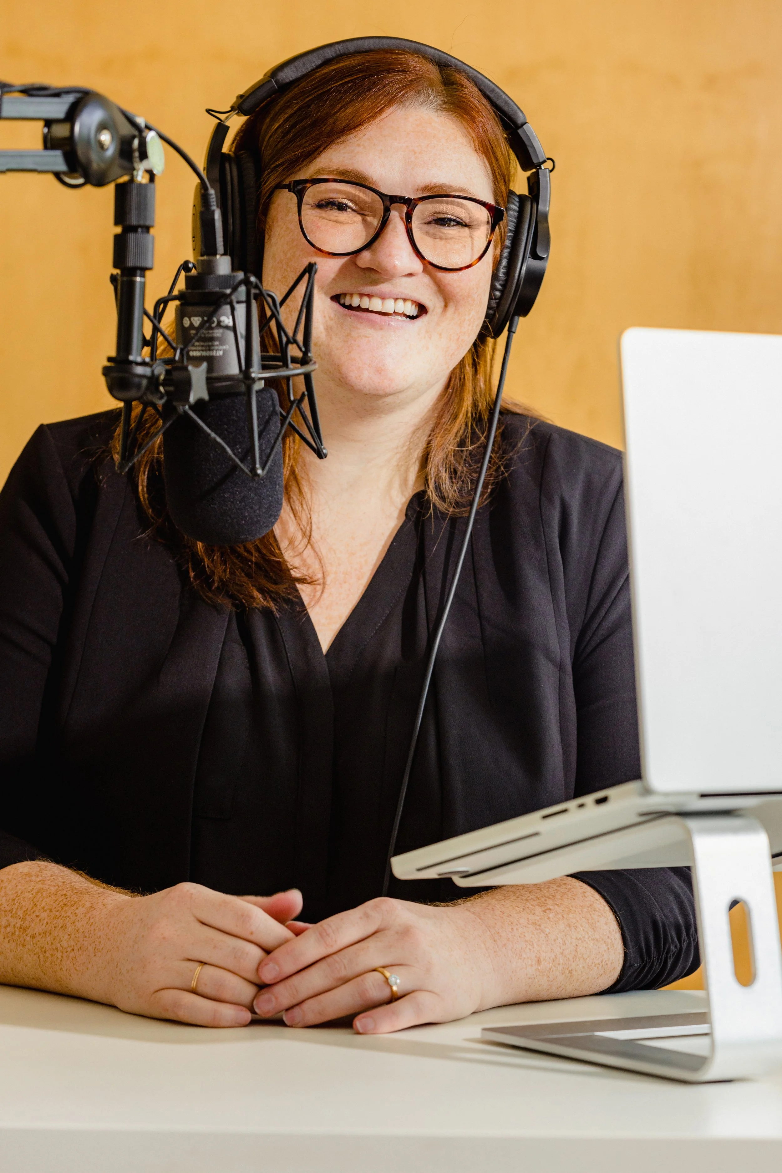 A woman with red hair and glasses smiling, sitting at a table with a laptop and a microphone in front of her, wearing headphones.