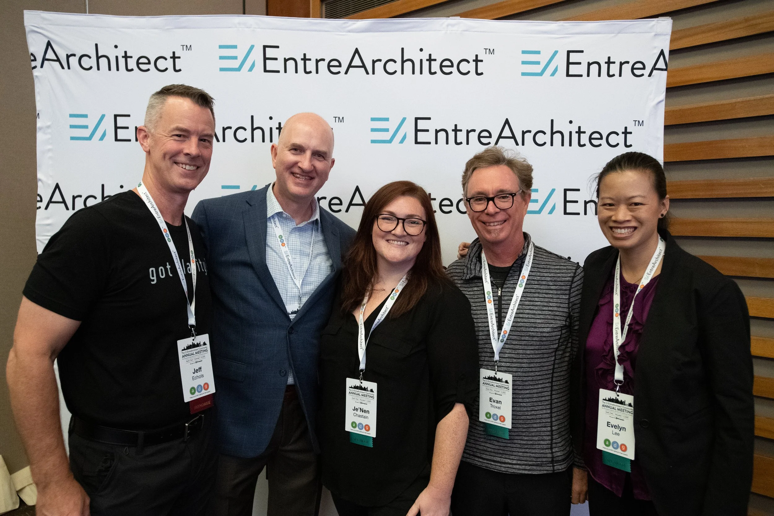Five people smiling and standing in front of a backdrop that reads 'EntreArchitect' at a conference or event. They are wearing name badges and lanyards.