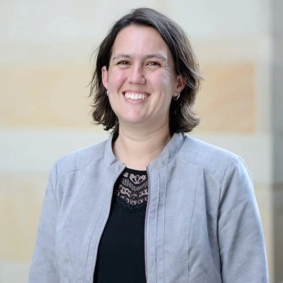 A smiling woman with short brown hair wearing a light gray jacket and black top, standing indoors against a blurred background.