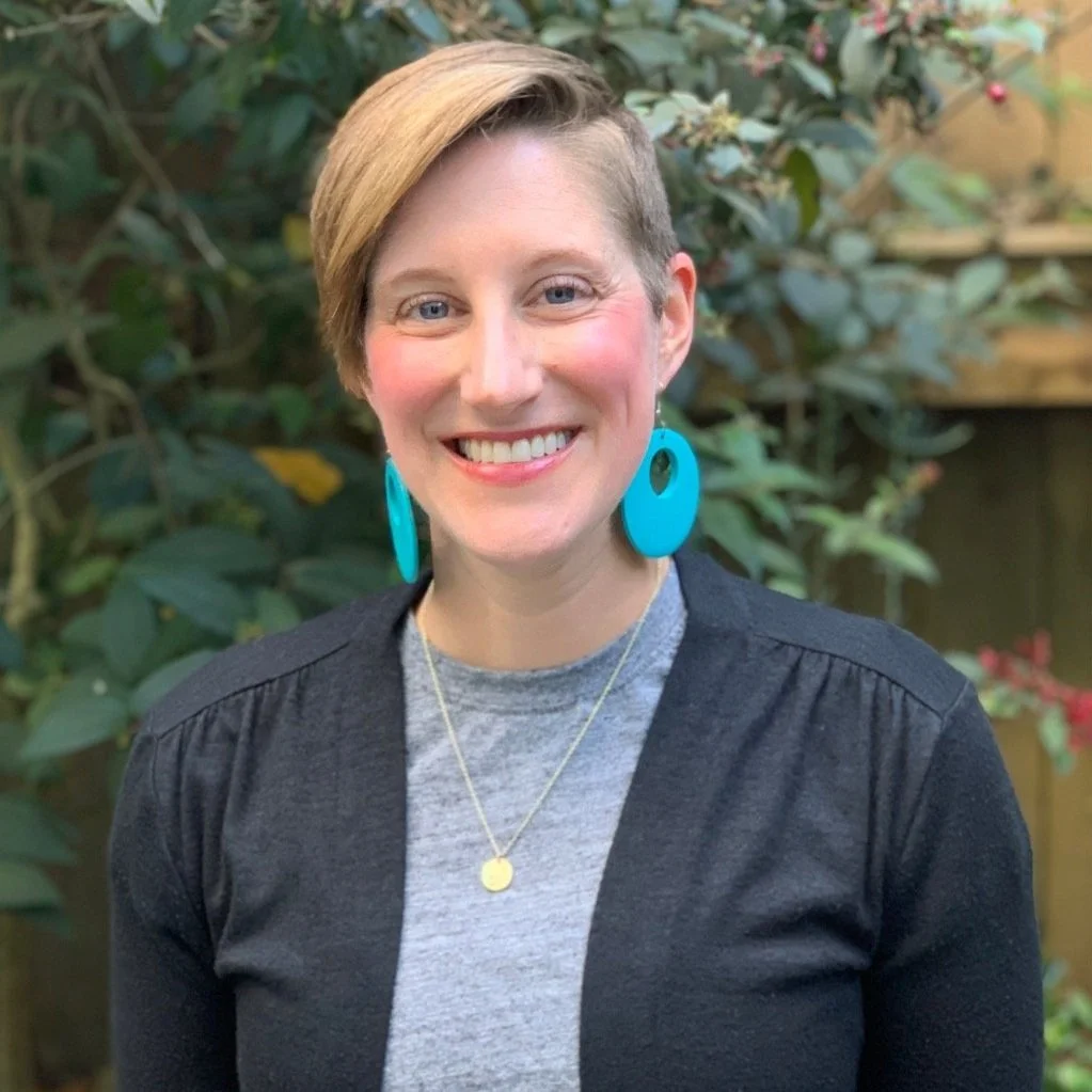 A smiling woman with short light brown hair wearing blue statement earrings, a grey shirt, a black cardigan, and a gold necklace, standing outdoors in front of green foliage.