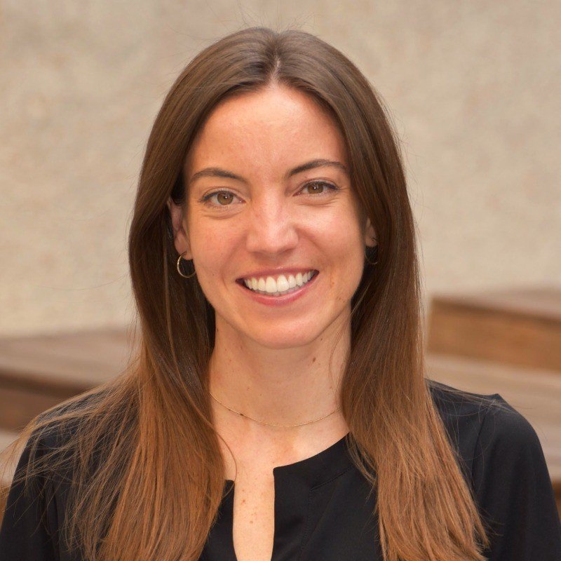 A woman with long brown hair, wearing earrings and a black top, smiling at the camera in an indoor setting.