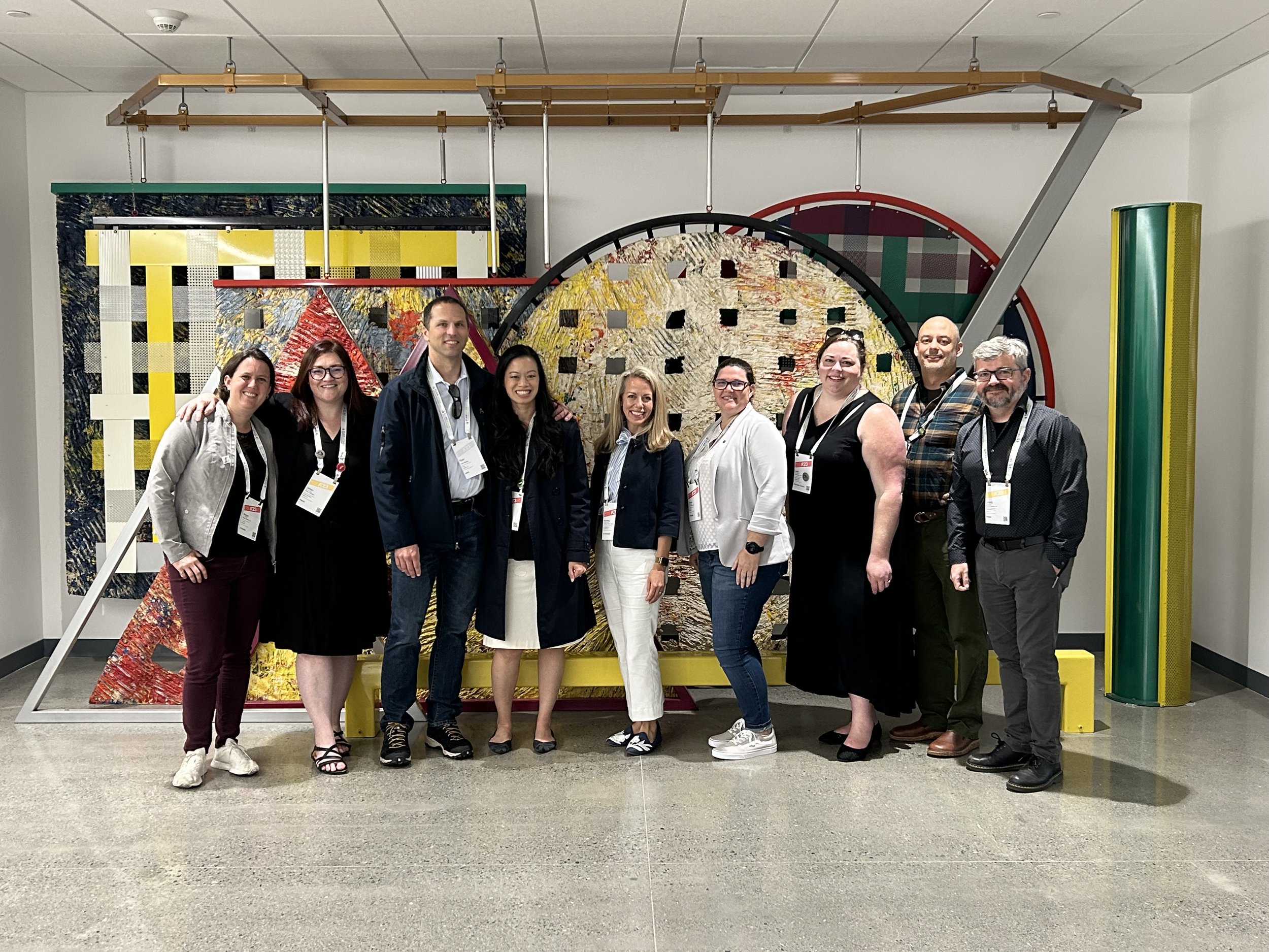 A group of ten diverse people standing together indoors in front of colorful abstract art and geometric sculpture. They are smiling and wearing conference name tags hanging from lanyards.