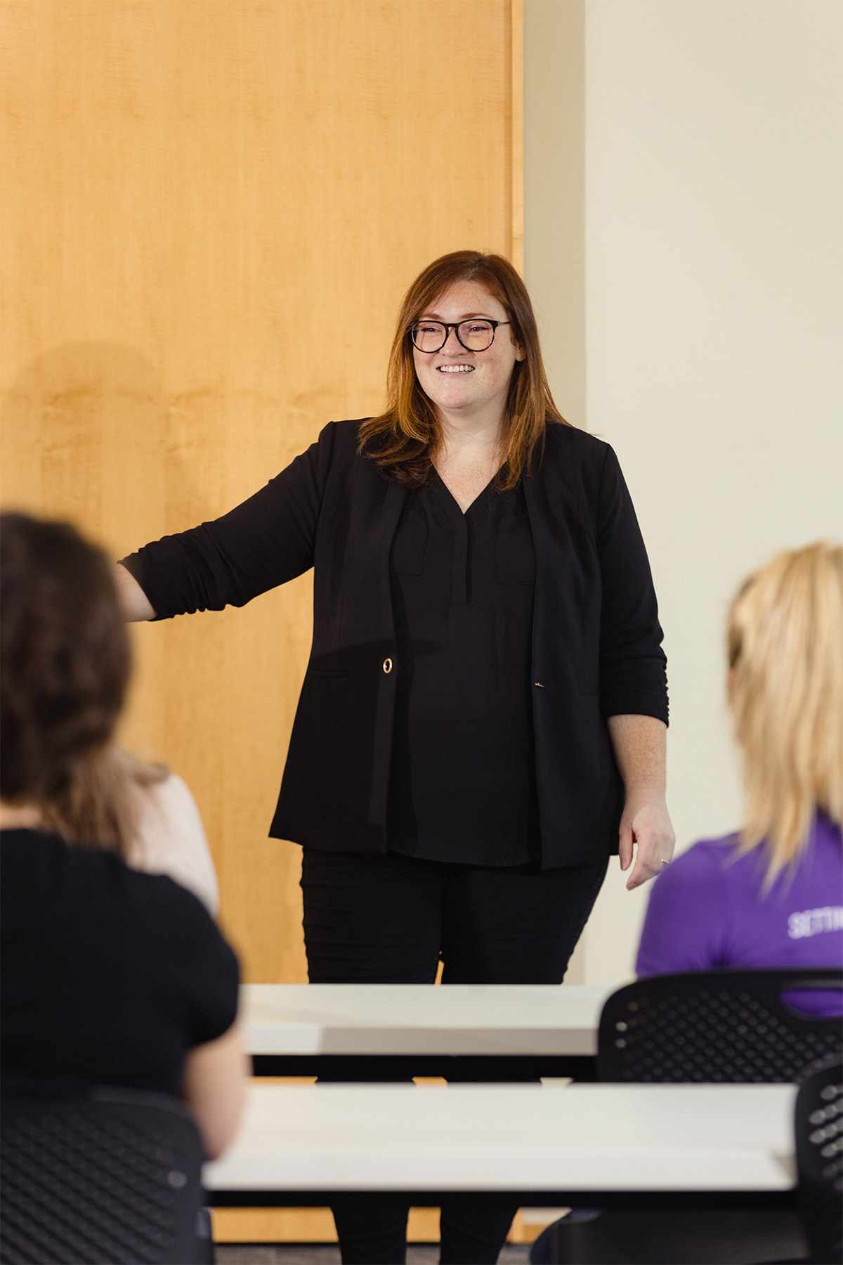 A woman with red hair, glasses, dressed in black, standing in front of a classroom giving a presentation to students.