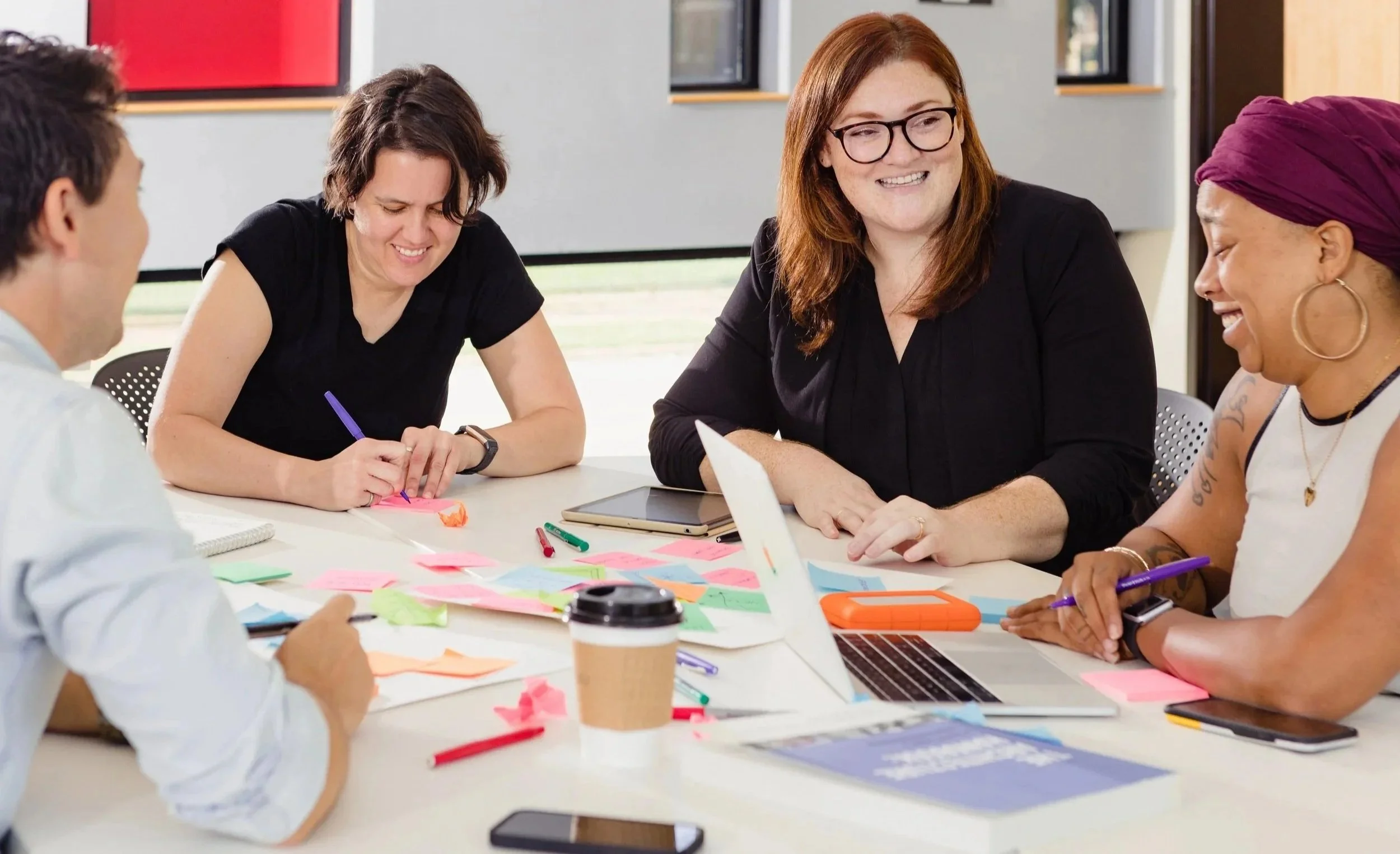Four women are sitting at a table engaged in a collaborative discussion or brainstorming session, with colorful sticky notes, markers, a laptop, and a disposable coffee cup on the table.