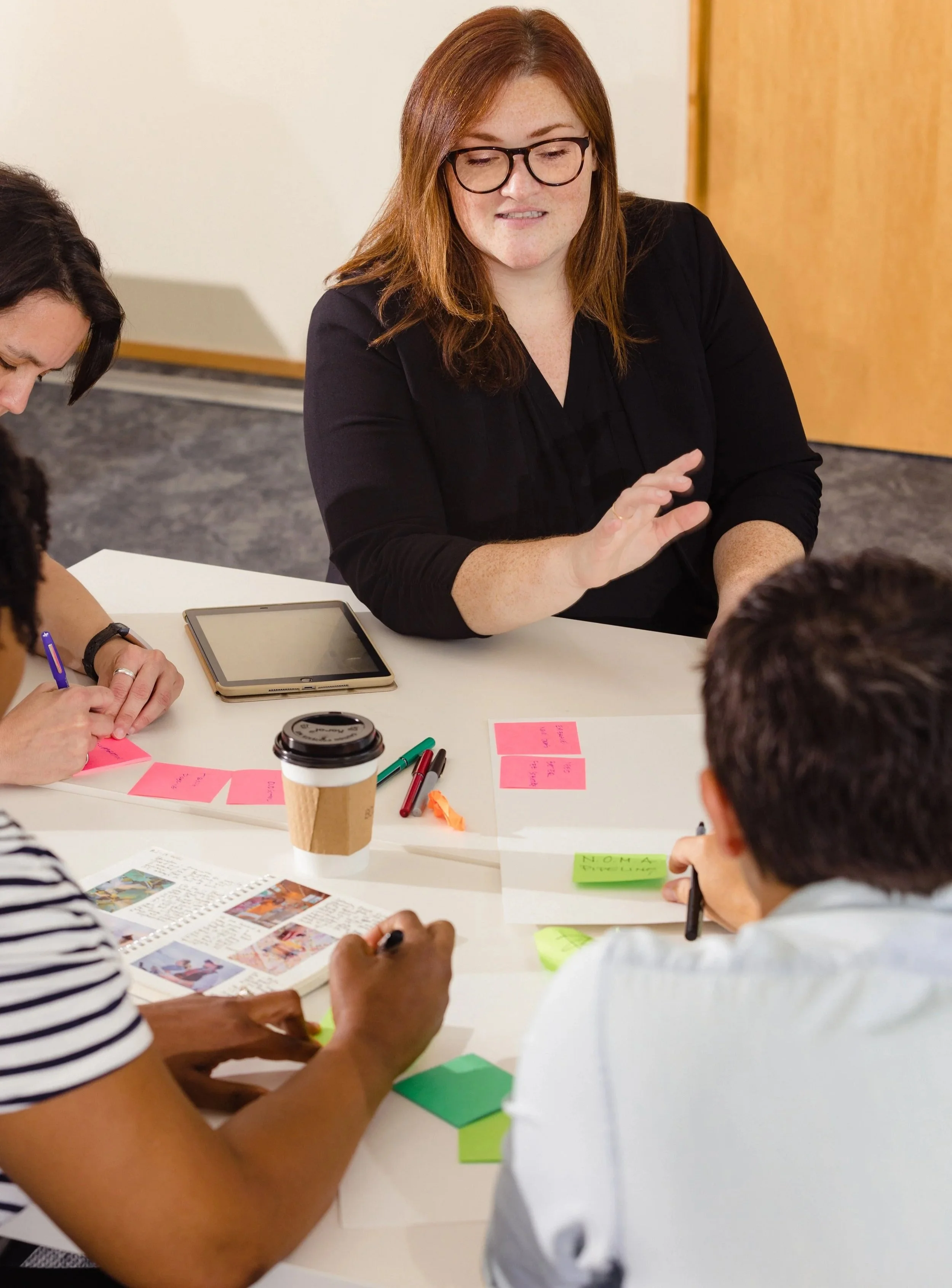 A group of diverse people sitting around a white table engaging in a discussion. A woman with red hair and glasses is speaking, with sticky notes, pens, a coffee cup, a tablet, and a notebook on the table.