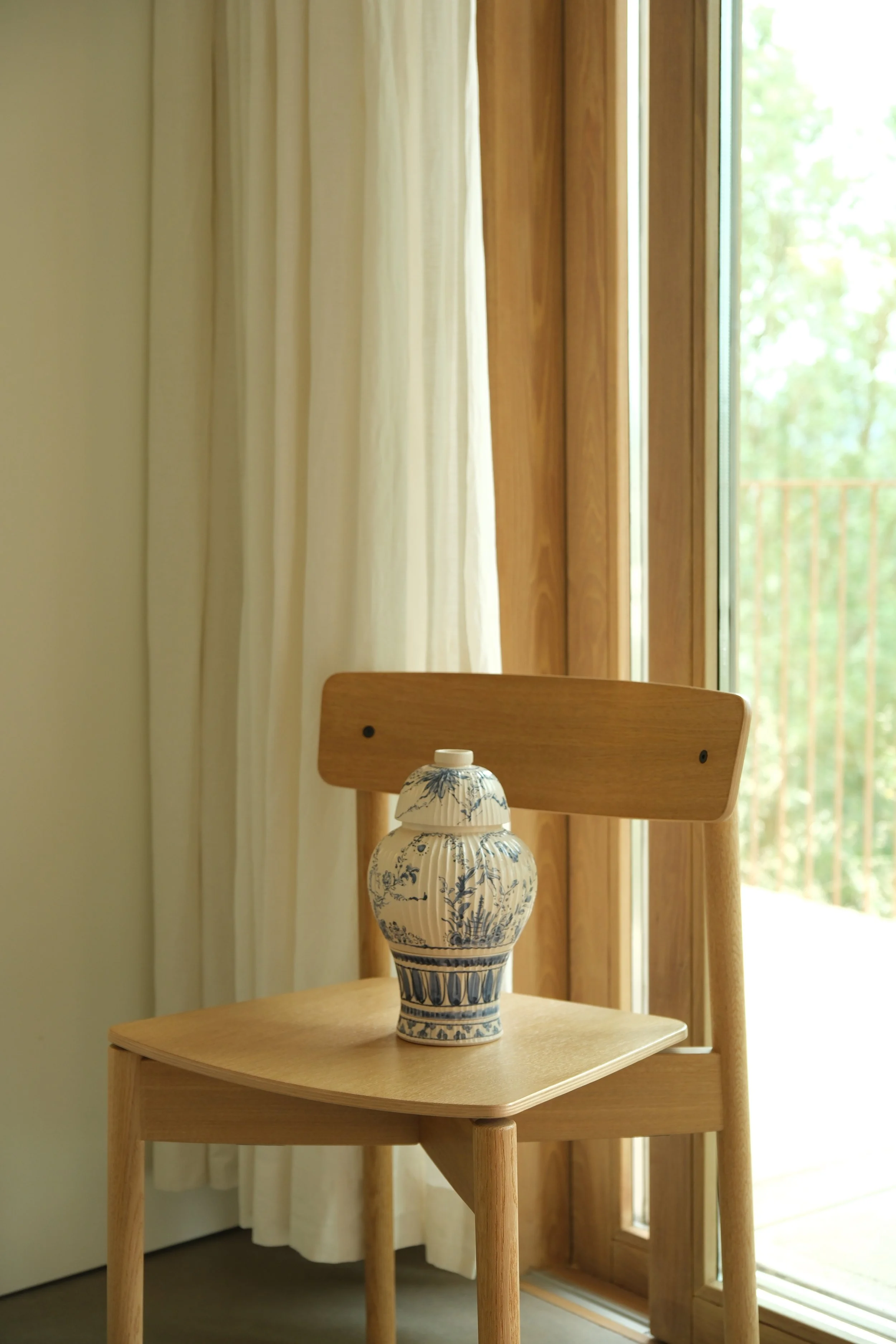 Vaso decorativo azul e branco em cima de uma mesa de madeira no interior próximo a uma janela de vidro com cortinas brancas ao lado.