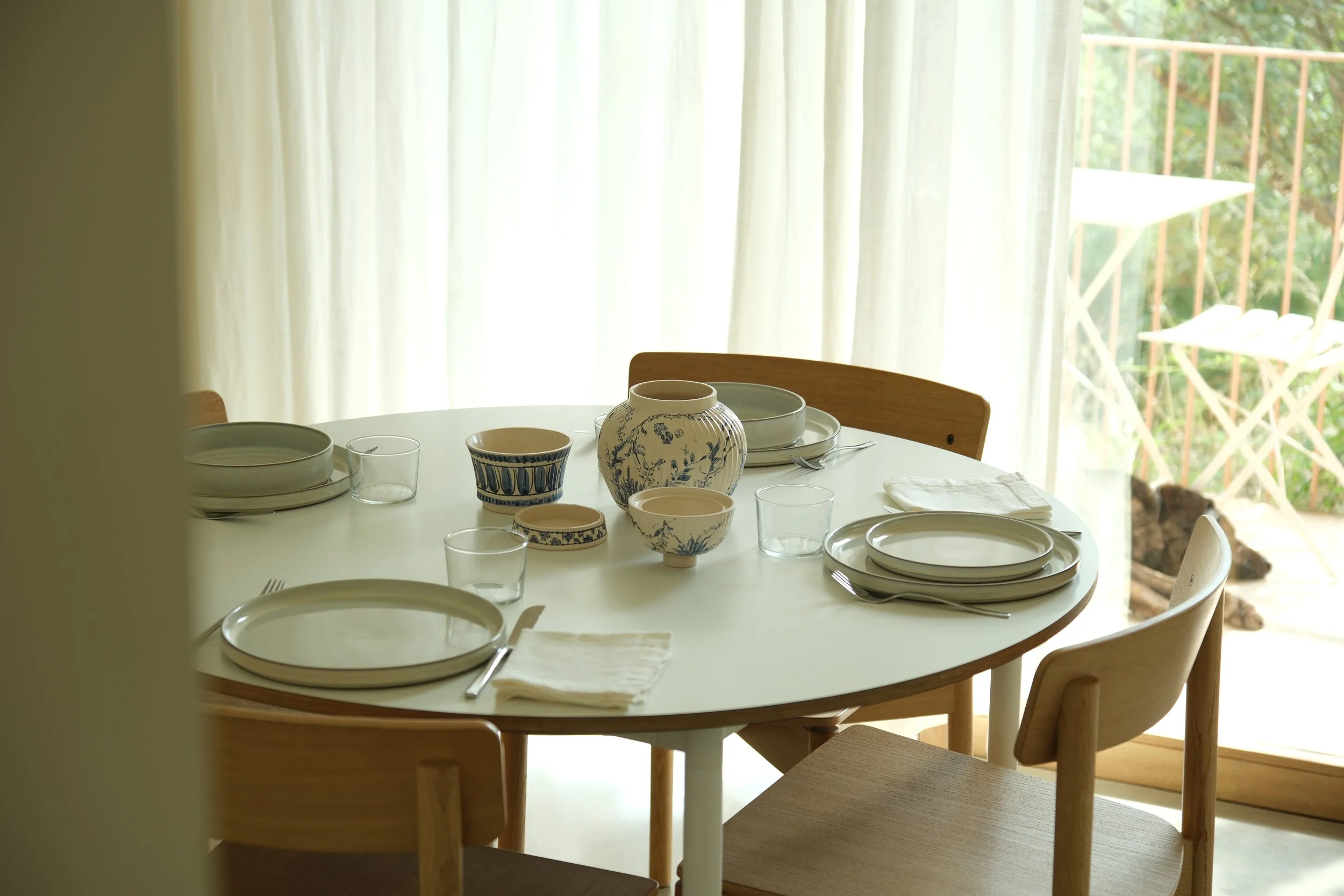 Mesa de jantar redonda com pratos, tigelas e copos de porcelana branca, cercada por cadeiras de madeira, próximo a uma janela com cortinas brancas e vista para um jardim com árvores.