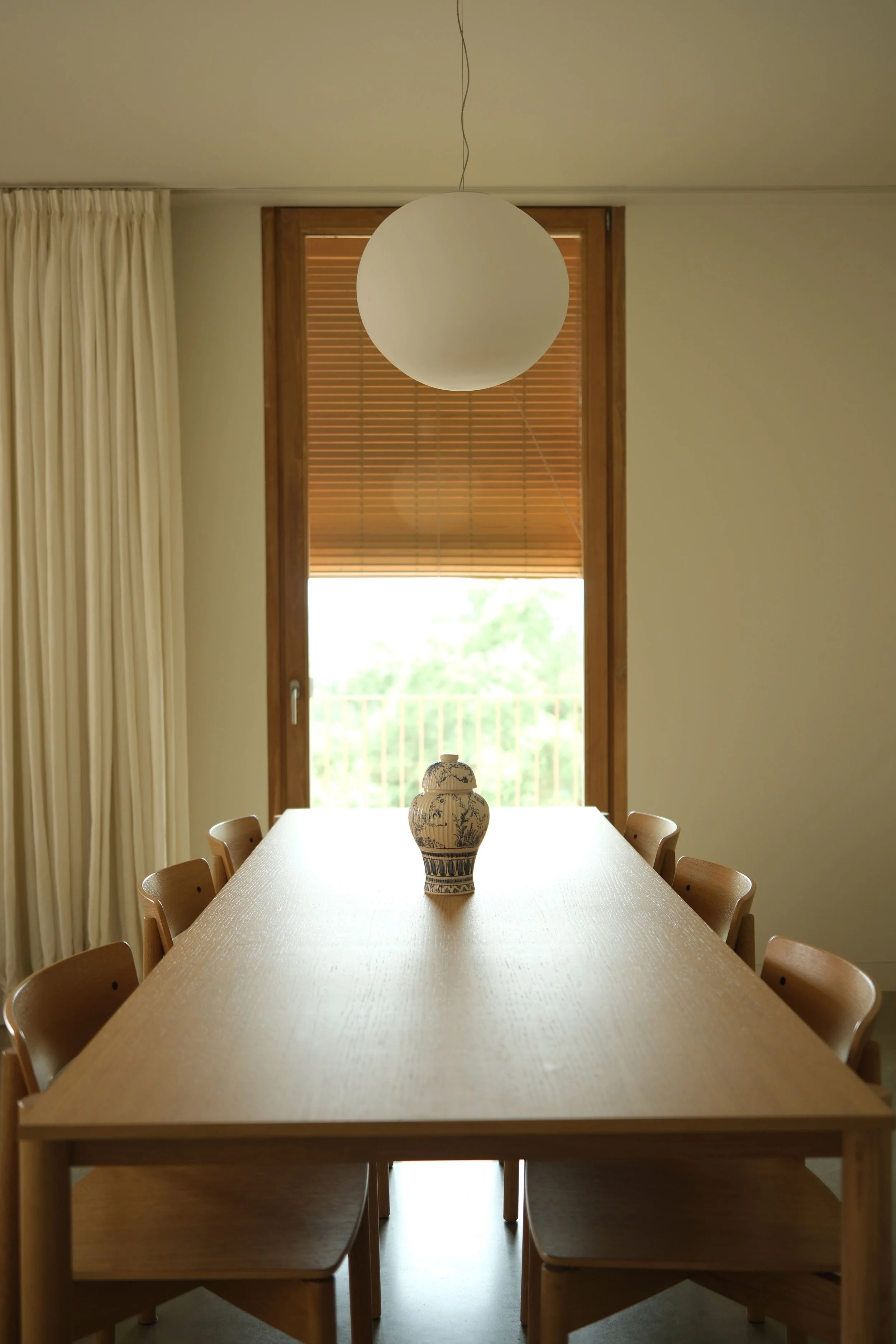 Sala de jantar com mesa de madeira comprida, ao fundo janela grande com cortinas brancas, persianas de madeira e candeeiro minimalista sobre a mesa. No centro da mesa, está a peça do projeto Há lugar para +1? da Oficina Feitiu destacada sobre a mesa.