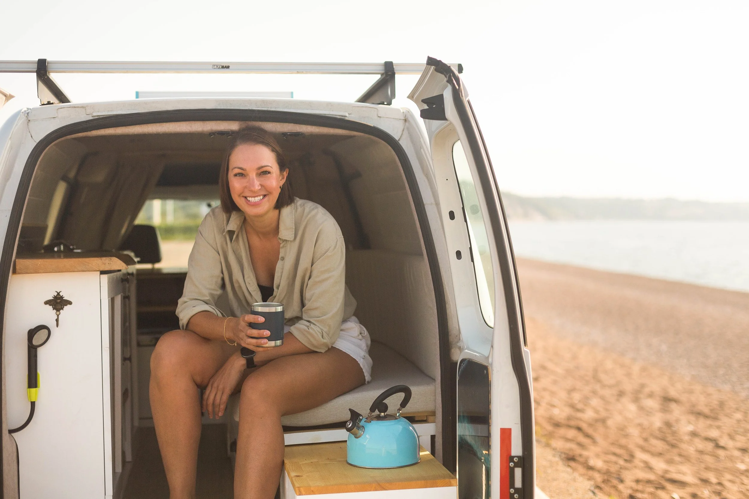 A woman sitting inside a camper van parked by a beach with a smile, holding a mug, with a kettle on a small wooden table nearby.