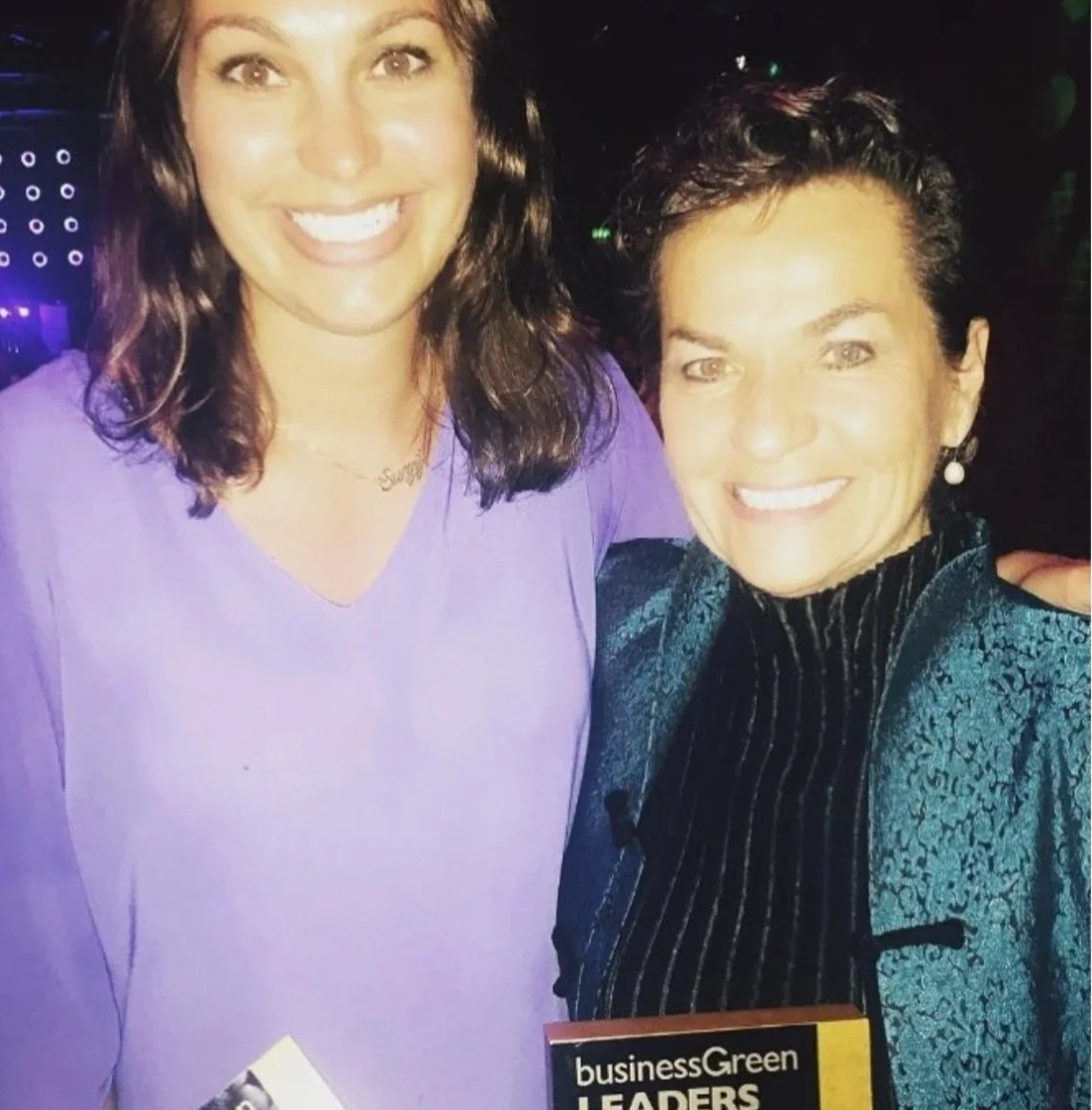 Suzy Greenwood and Christiana Figueres. Two women smiling for a photo at a social event, one wearing a purple top and the other in a textured jacket, both appearing happy and friendly.