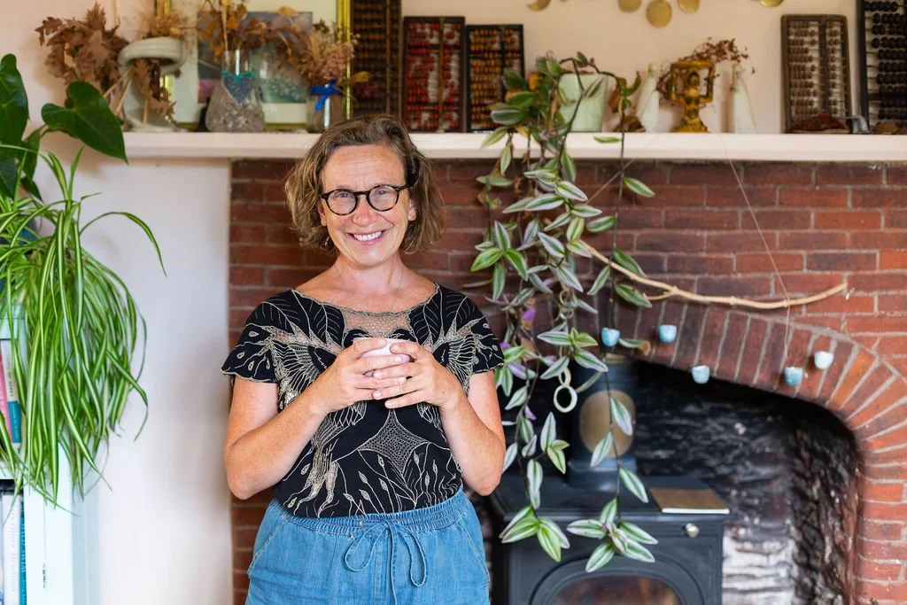 A smiling woman with glasses and curly hair standing in front of a brick fireplace, holding a cup. She is surrounded by green plants and decorative items on the mantel.
